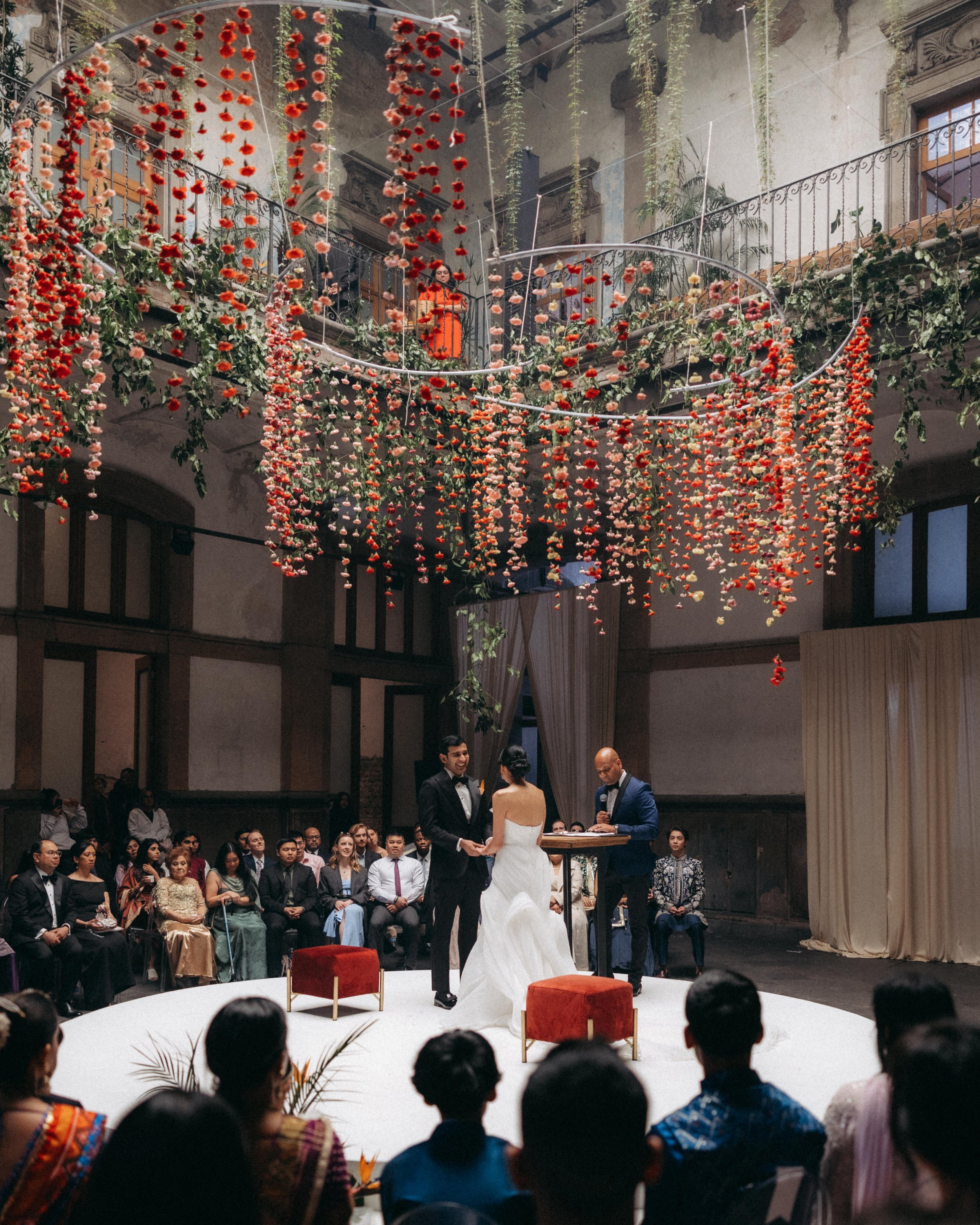 A wedding ceremony taking place indoors with a bride and groom holding hands, standing in front of an officiant at a small table. The ceremony is surrounded by seated guests and decorated with hanging red and pink flowers and green foliage above. The