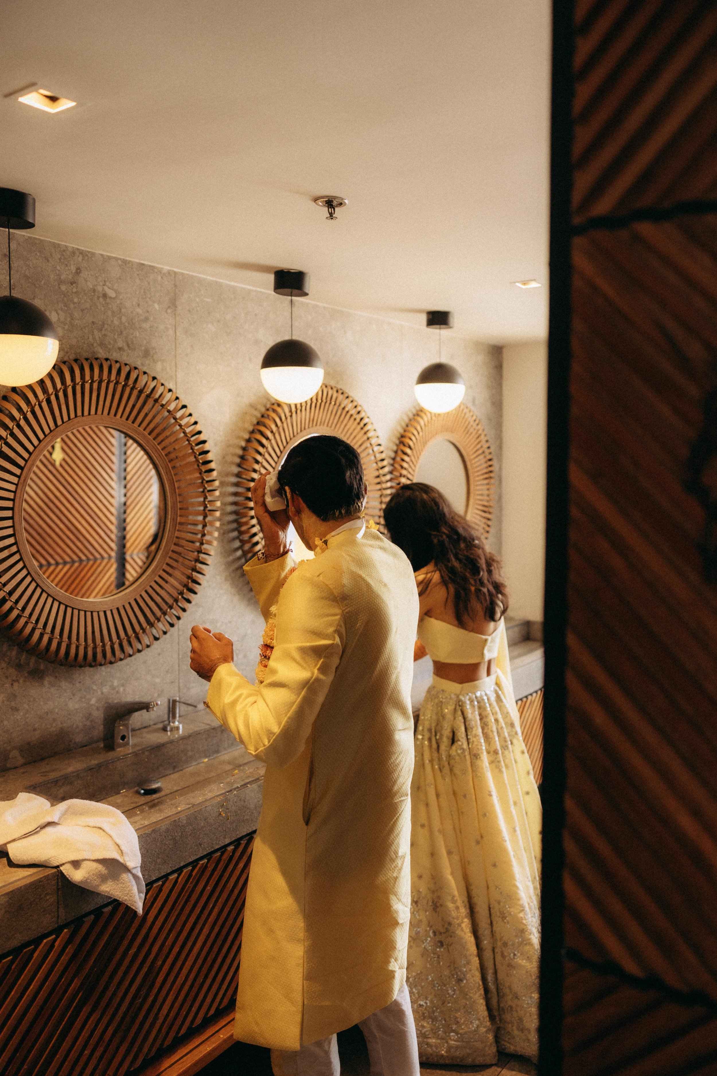 People wearing traditional Indian attire in front of decorative mirrors and hanging lights in a room.