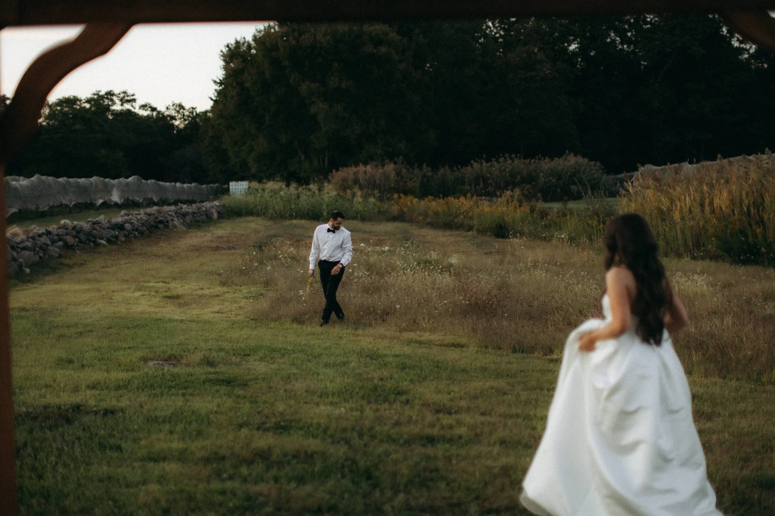 A couple dressed in formal wear, a man in a tuxedo and a woman in a white dress, are outdoors in a grassy field during dusk or early evening, with trees and a stone wall in the background.