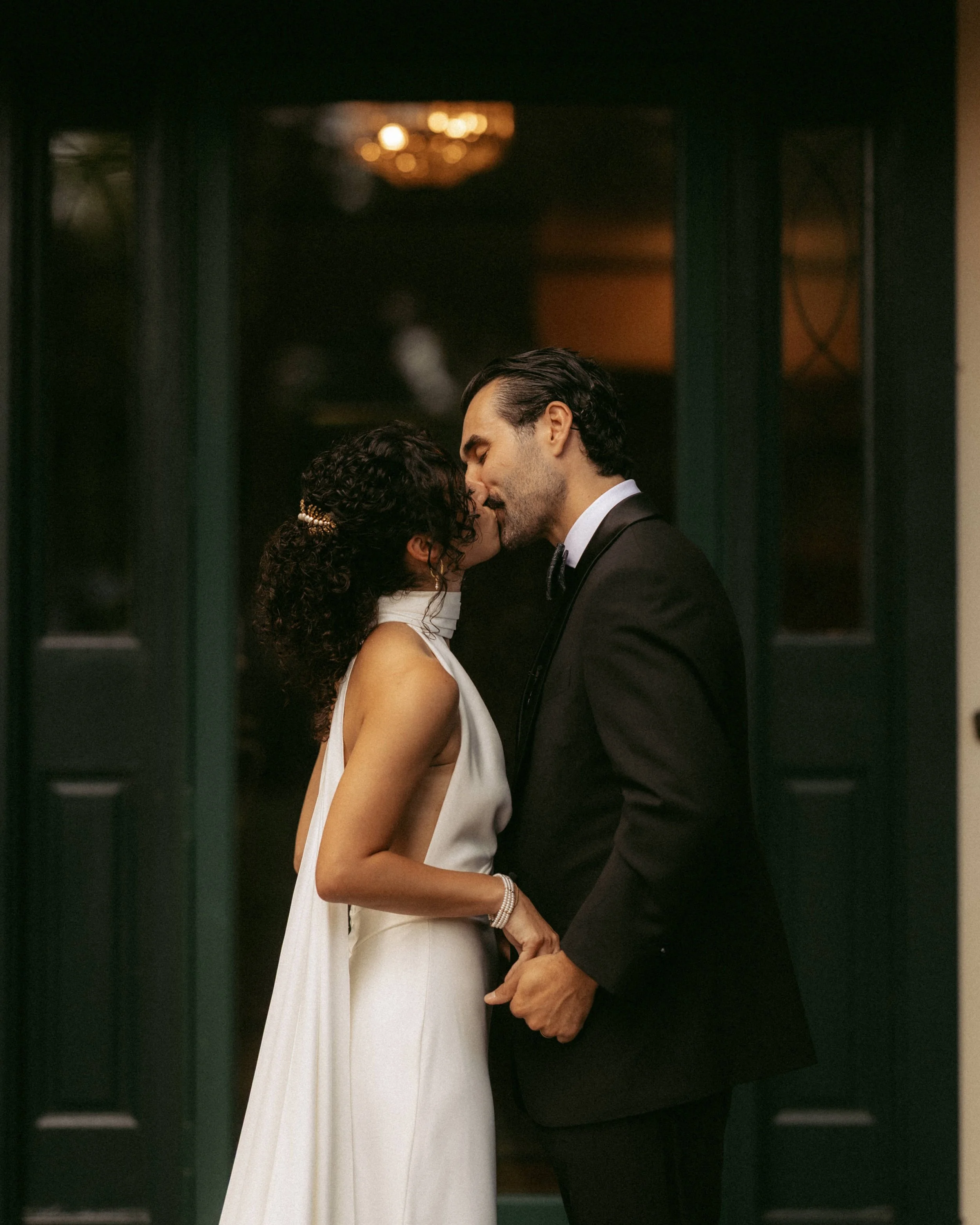 A man and woman are sharing a kiss in formal attire, standing close together in a dimly lit setting.