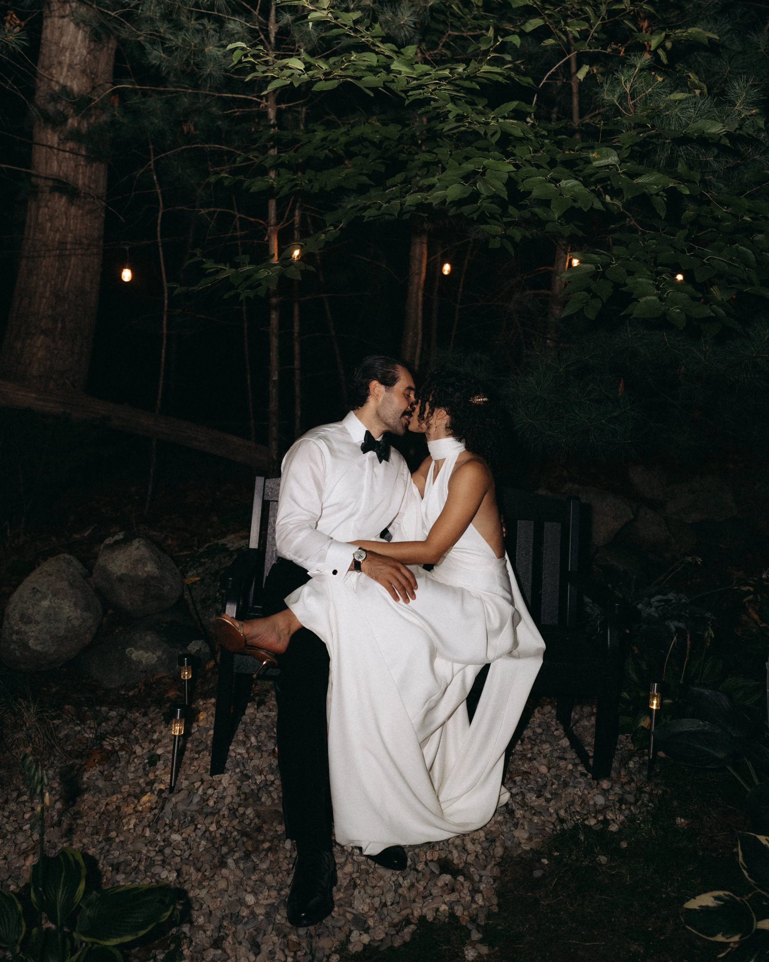 A couple dressed formally, with the man in a white shirt and black bow tie and the woman in a white dress, sitting closely and kissing outdoors at night with string lights hanging above and small lights on the ground.