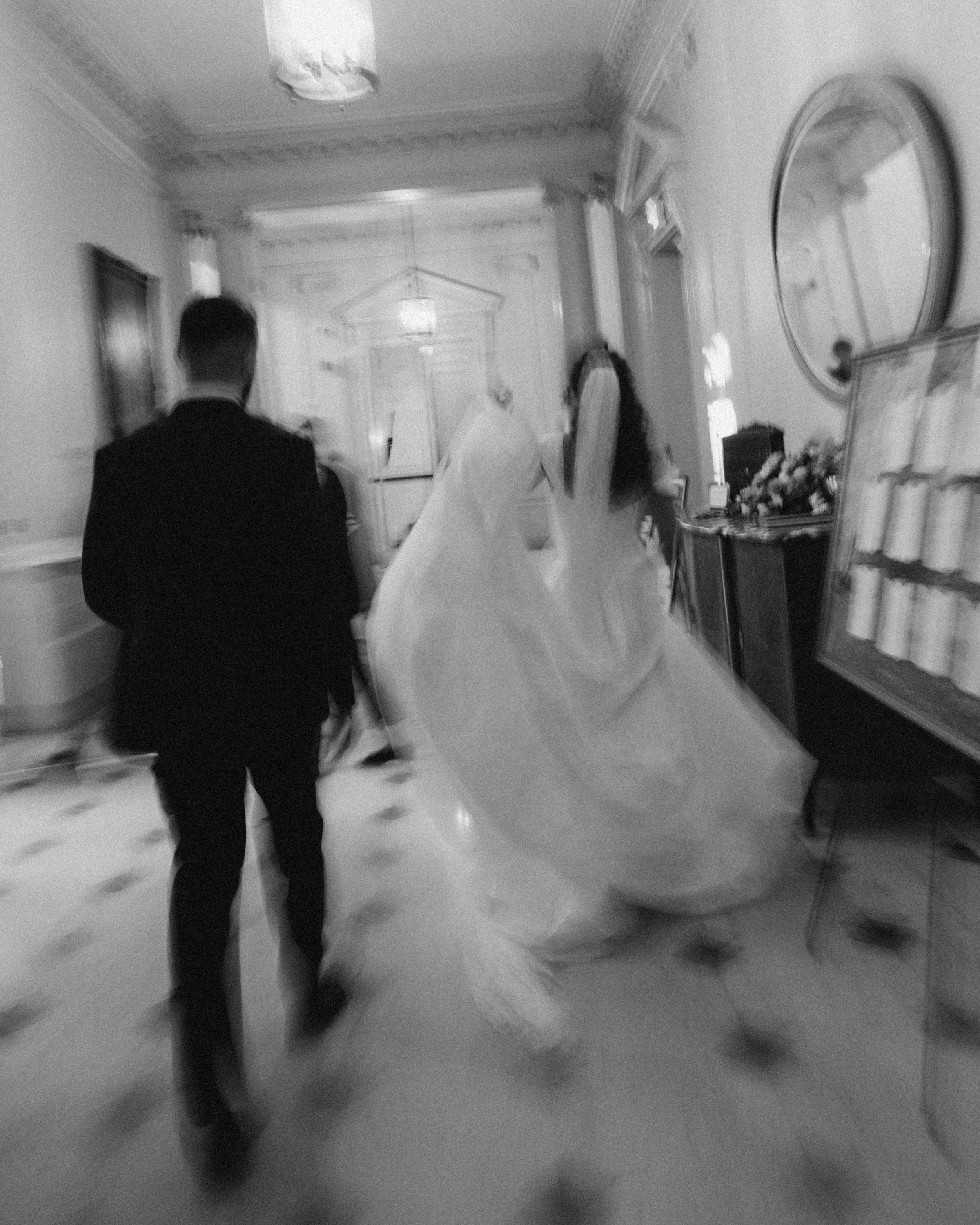 A person in a suit walking through an elegant, vintage-style room with ornate crown molding, a large oval mirror, and floral arrangements.
