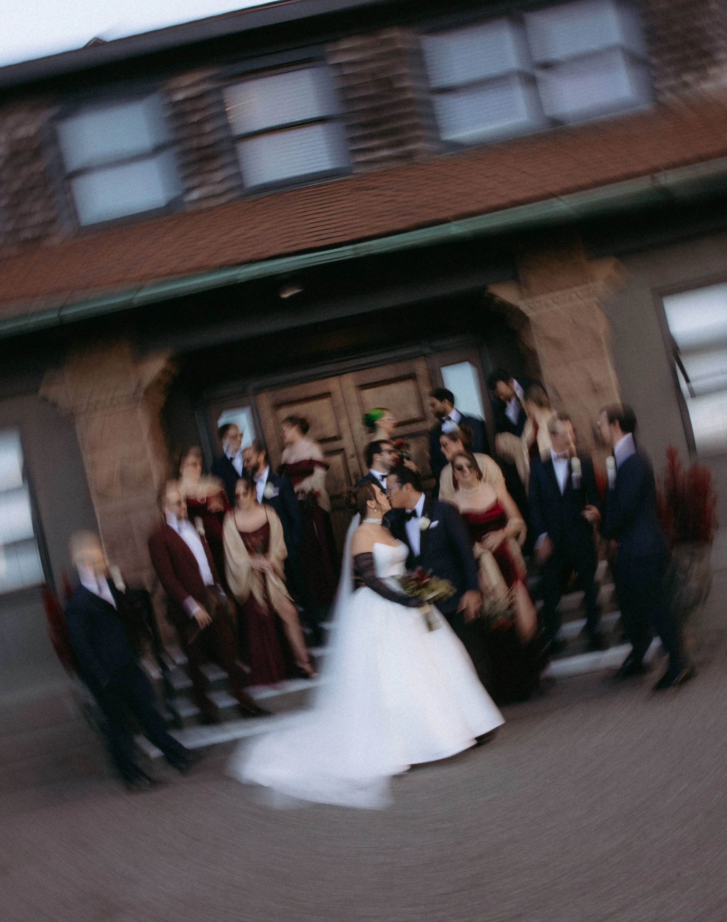 A group of people dressed formally, possibly at a wedding or formal event, standing outside a brick building with a large door, some engaged in conversation.