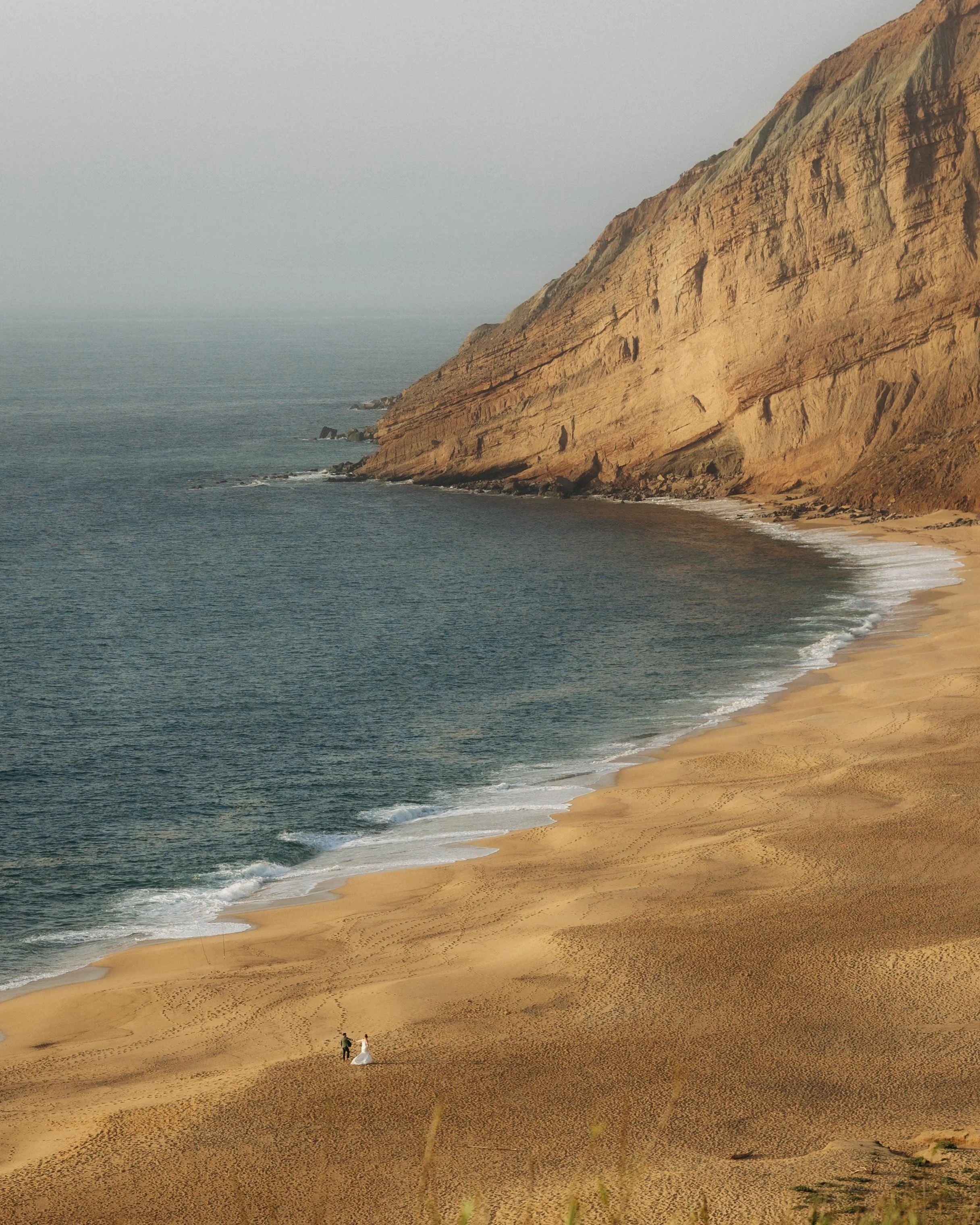 A couple, possibly a bride and groom, walking along a sandy beach at the base of a large, brown rocky cliff during daylight hours.