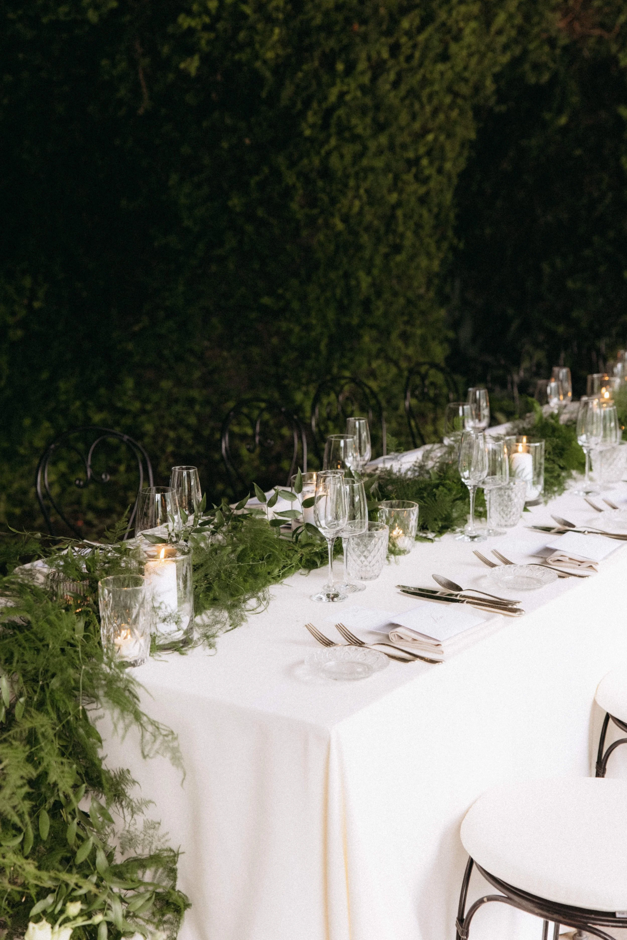 A long banquet table decorated with greenery, candles, wine glasses, and white napkins, set outdoors at night with trees in the background.