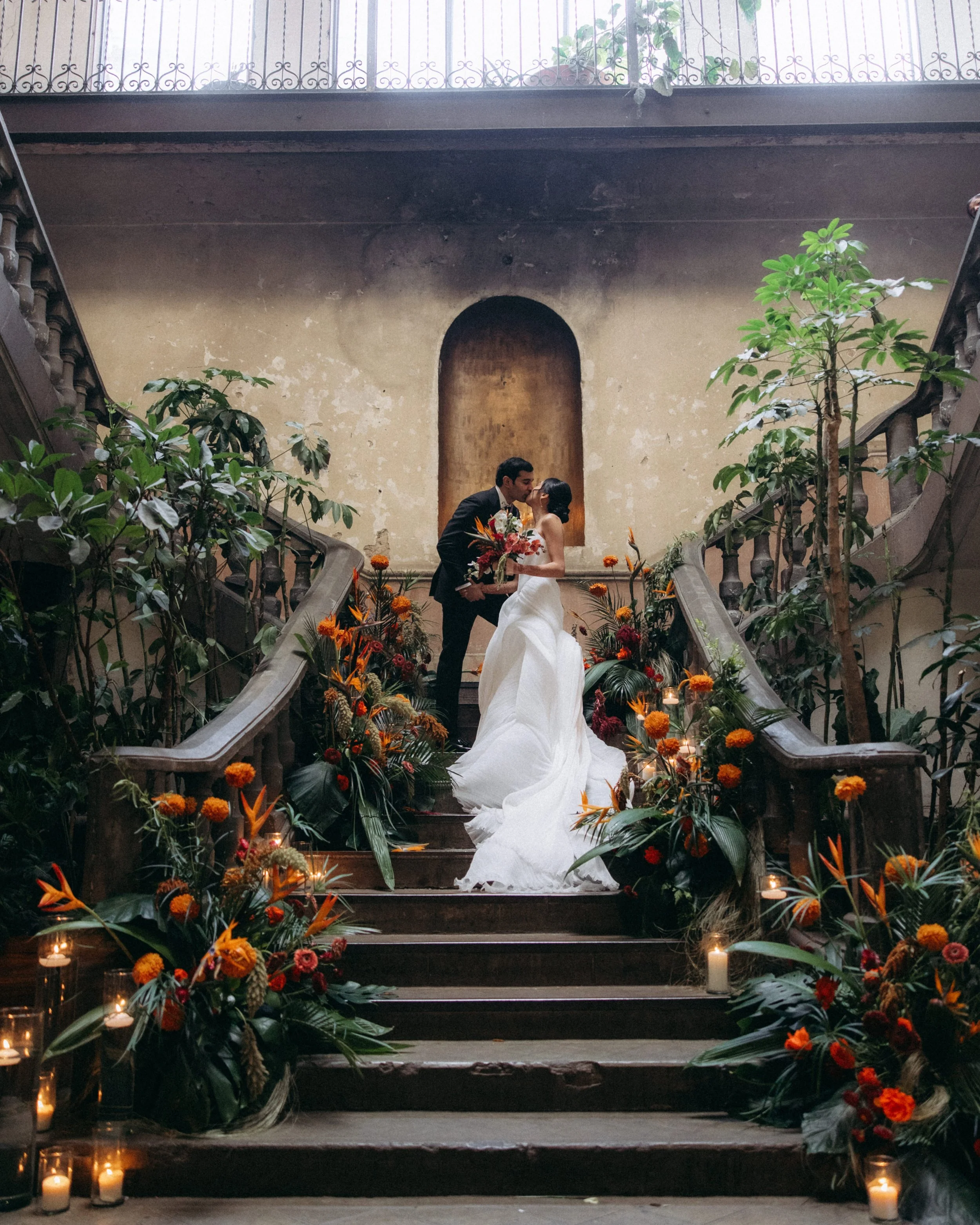 A bride and groom sharing a kiss on a staircase decorated with colorful flowers and candles, inside a building with a high ceiling and a balcony with a wrought iron railing.