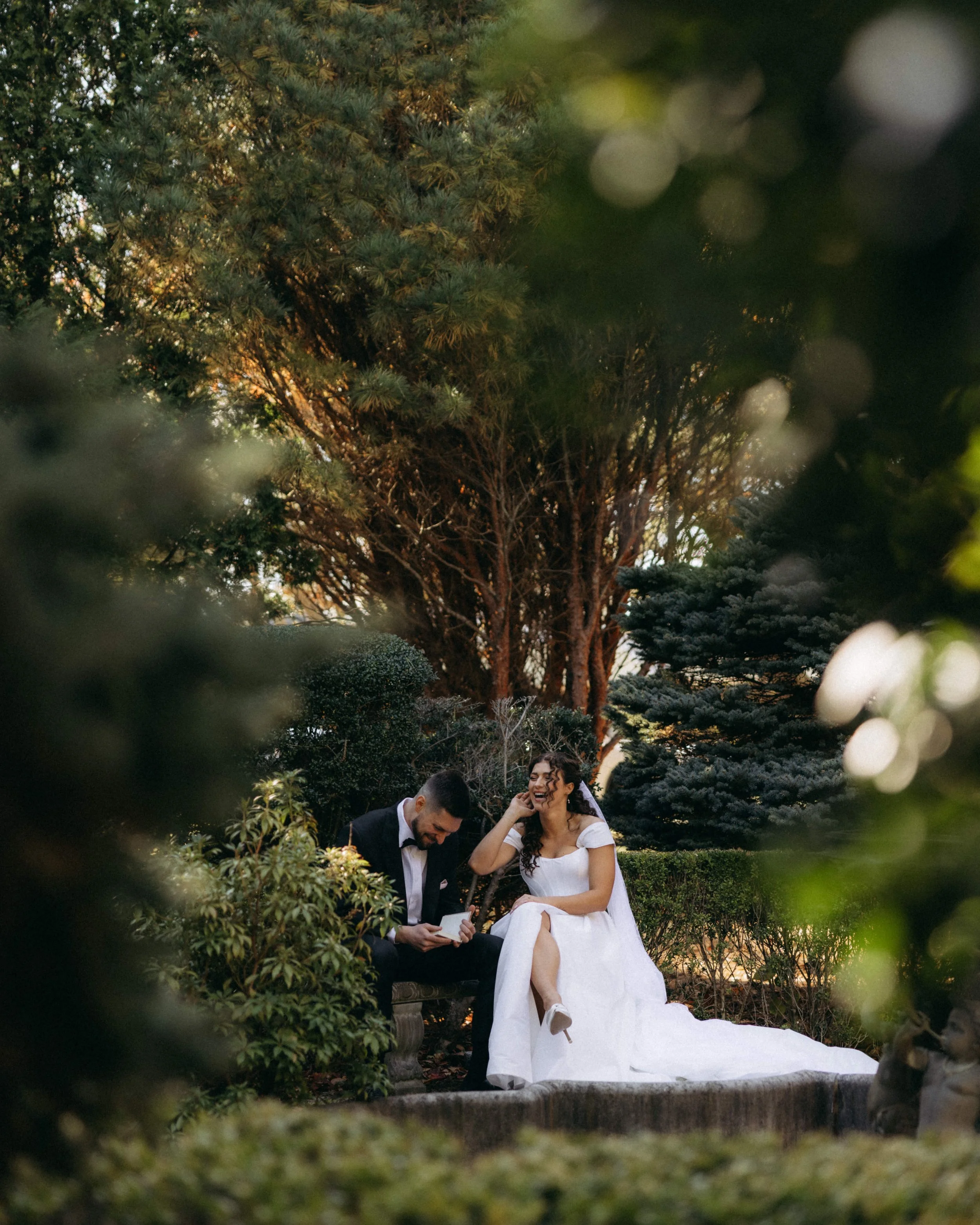 A bride and groom sitting together outdoors, surrounded by trees and shrubs, with the bride laughing and smiling, the groom looking at a piece of paper, possibly a wedding vow or speech.