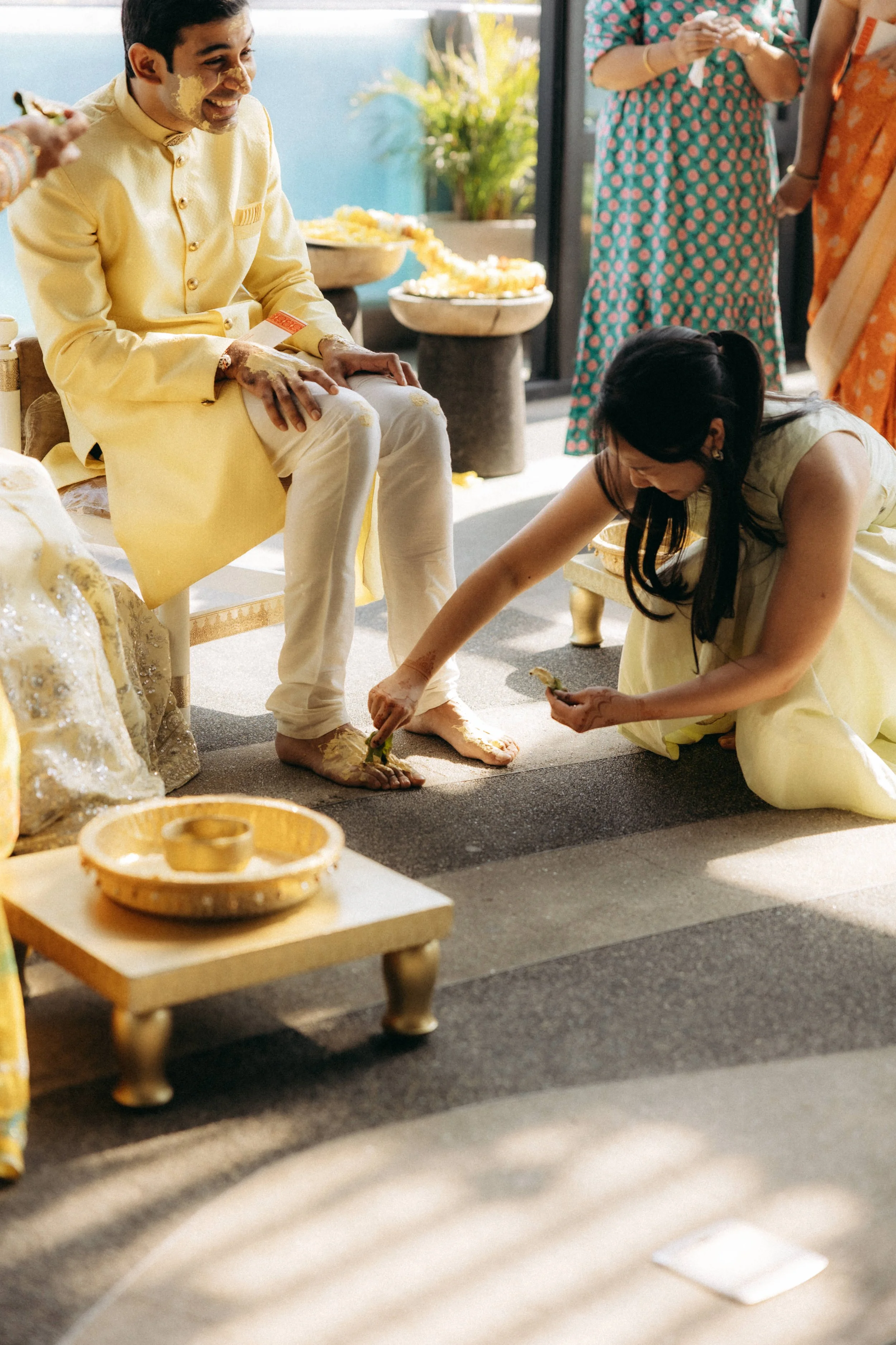 A man in traditional yellow Indian attire is laughing while sitting on a decorated chair, with a woman kneeling on the floor in front of him, applying turmeric and other substances to his feet during a celebration. Several women are standing in the b
