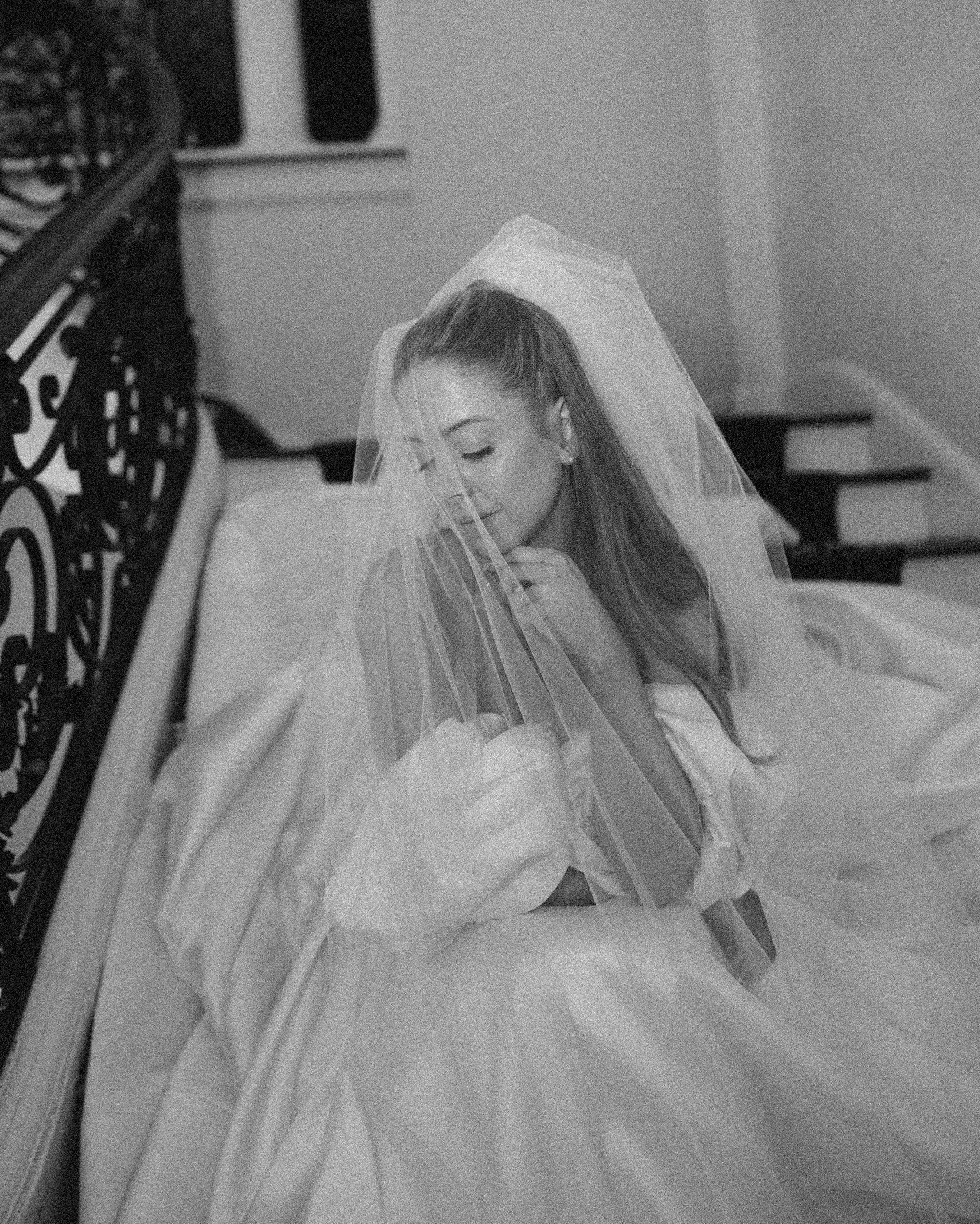 A woman with long hair and pearl earrings sitting at a piano with a veil over her head, smiling, inside a house with stairs in the background, black and white photo.