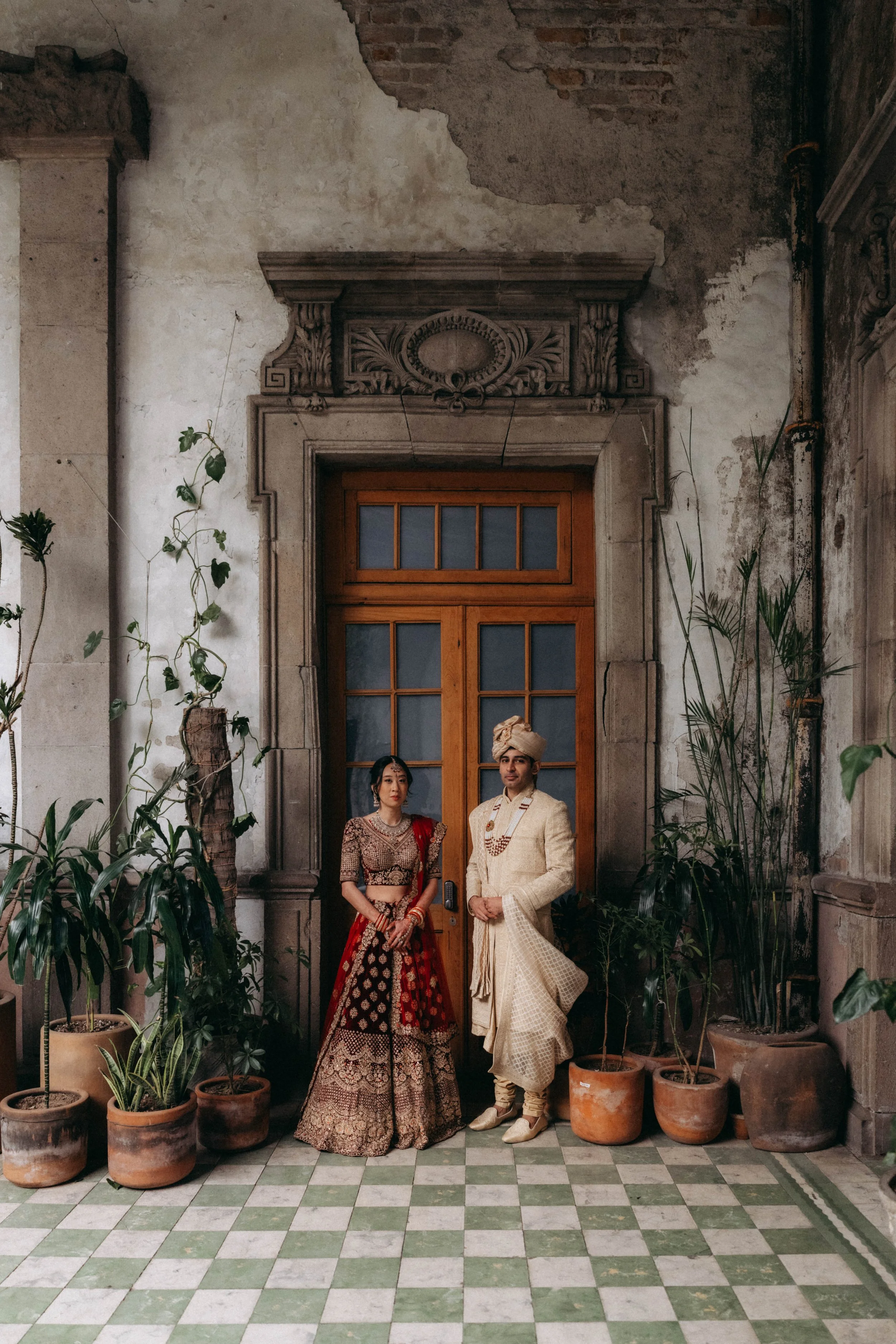 A couple dressed in traditional Indian wedding attire standing in front of a rustic wooden door with stone and weathered wall, surrounded by potted plants on a checkered tile floor.