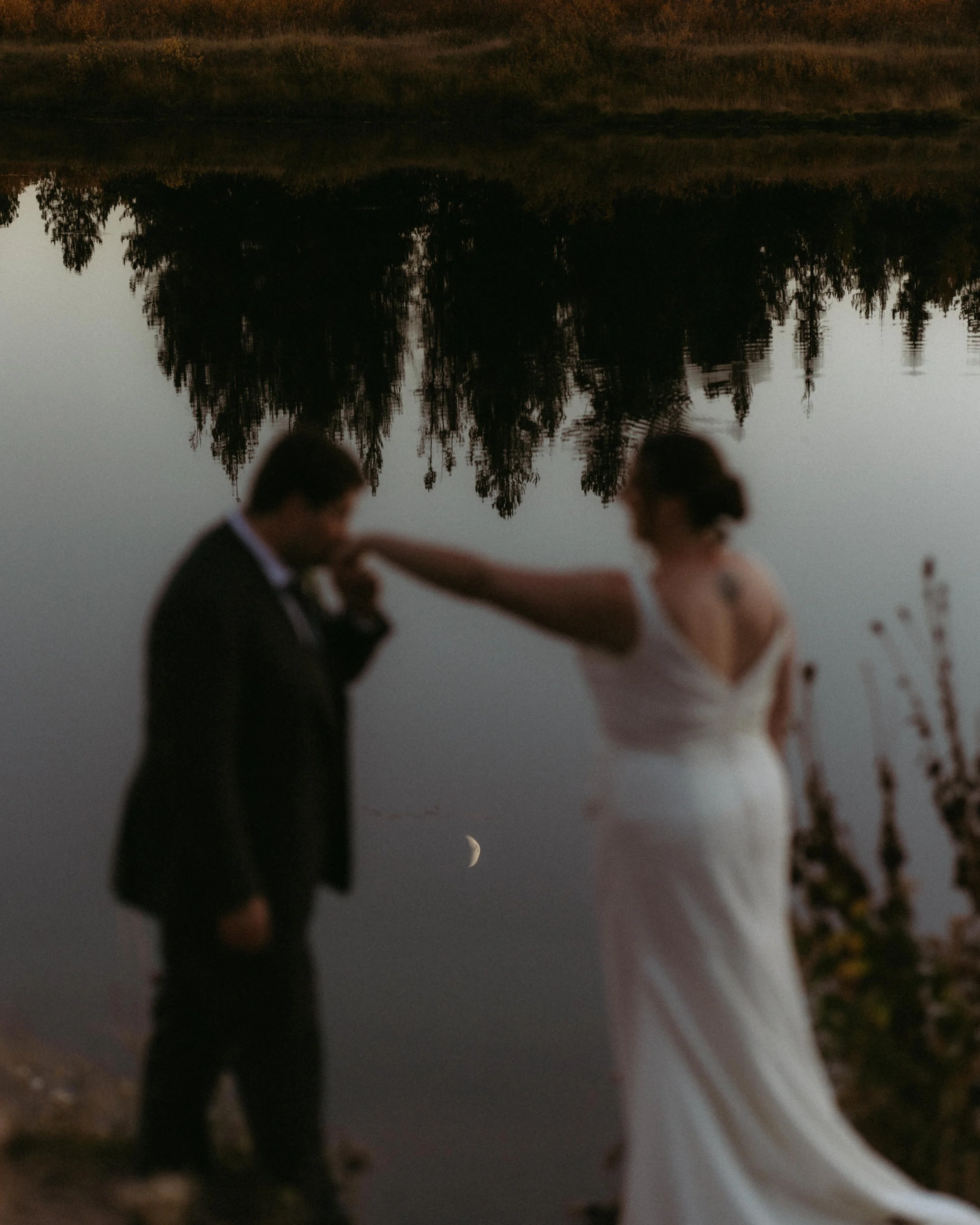 A man in a tuxedo and a woman in a white gown are holding hands and dancing on a beach near a body of water with reflected trees in the background at dusk.