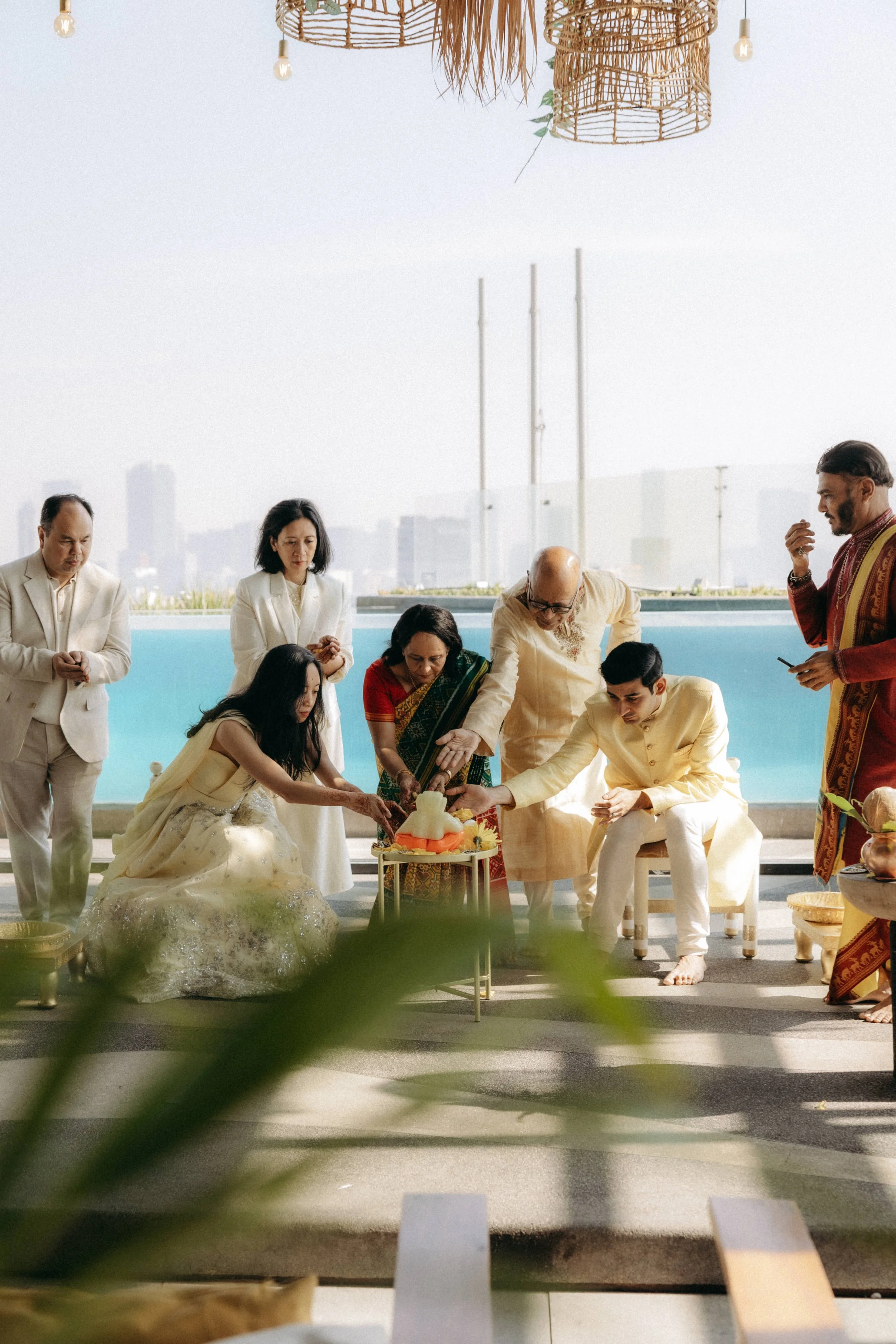 A group of people dressed in traditional and formal attire celebrating a special occasion on a terrace with city skyline in the background, with some participants cutting a cake. The scene includes hanging woven light fixtures and decorative plants.