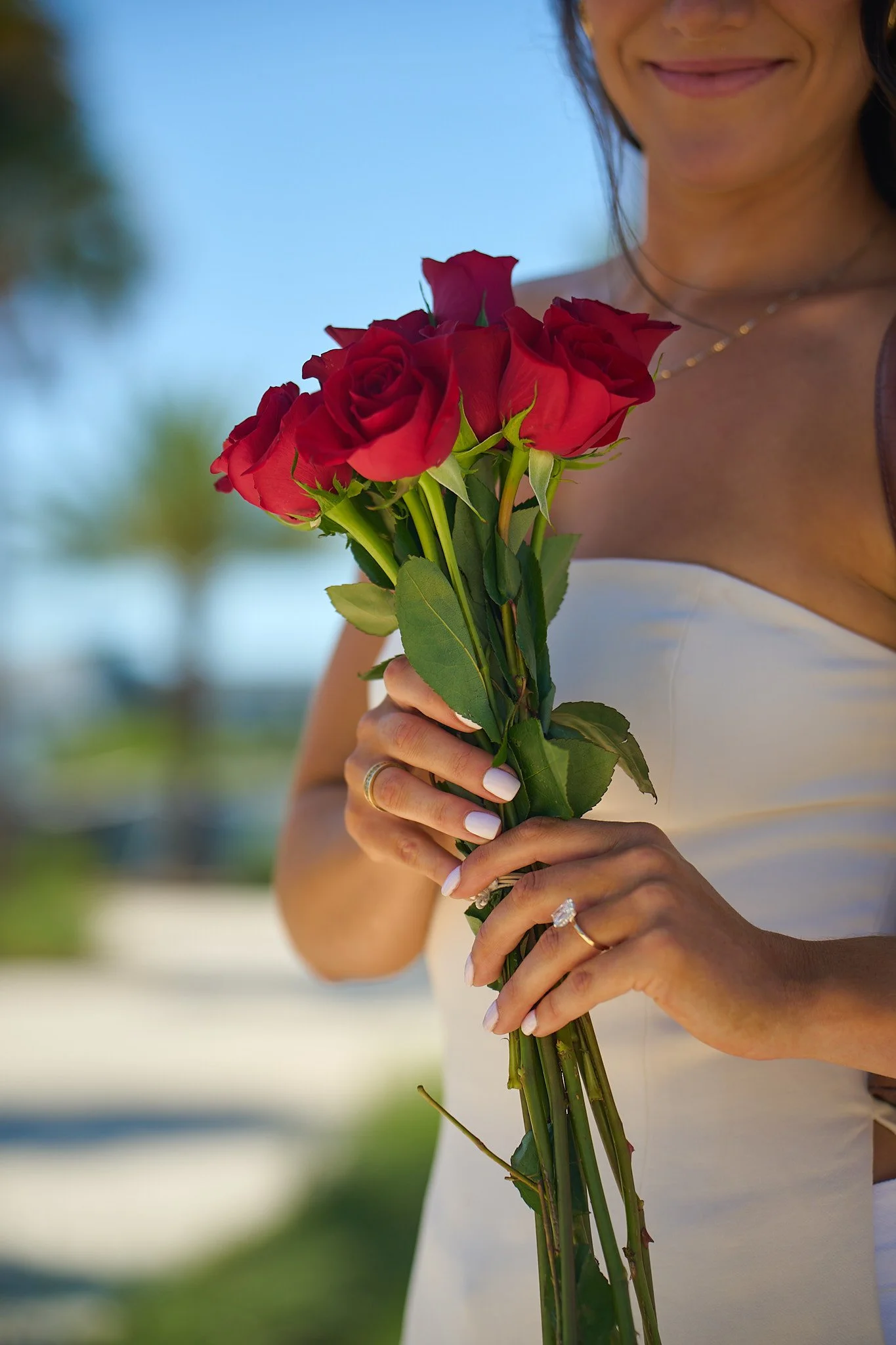 A woman in a white strapless dress holding a bouquet of red roses outdoors, smiling slightly, with blurred green trees and blue sky in the background.