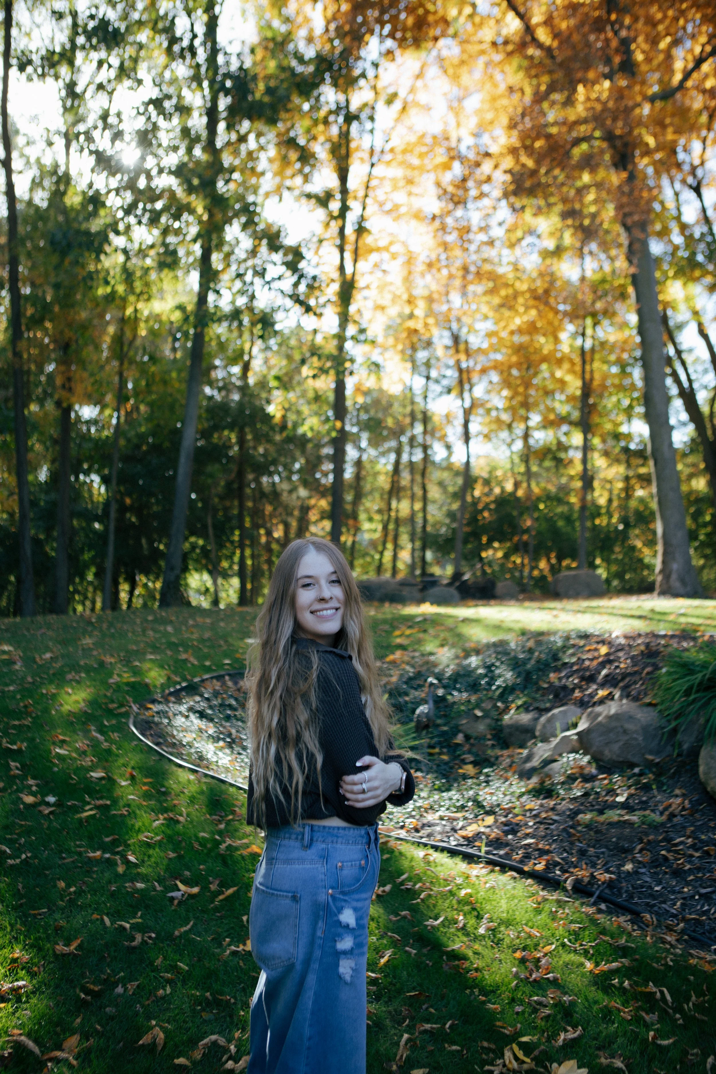 A young woman with long wavy hair smiling in a park during autumn, surrounded by trees with colorful leaves, standing on grass near a curved small stream.