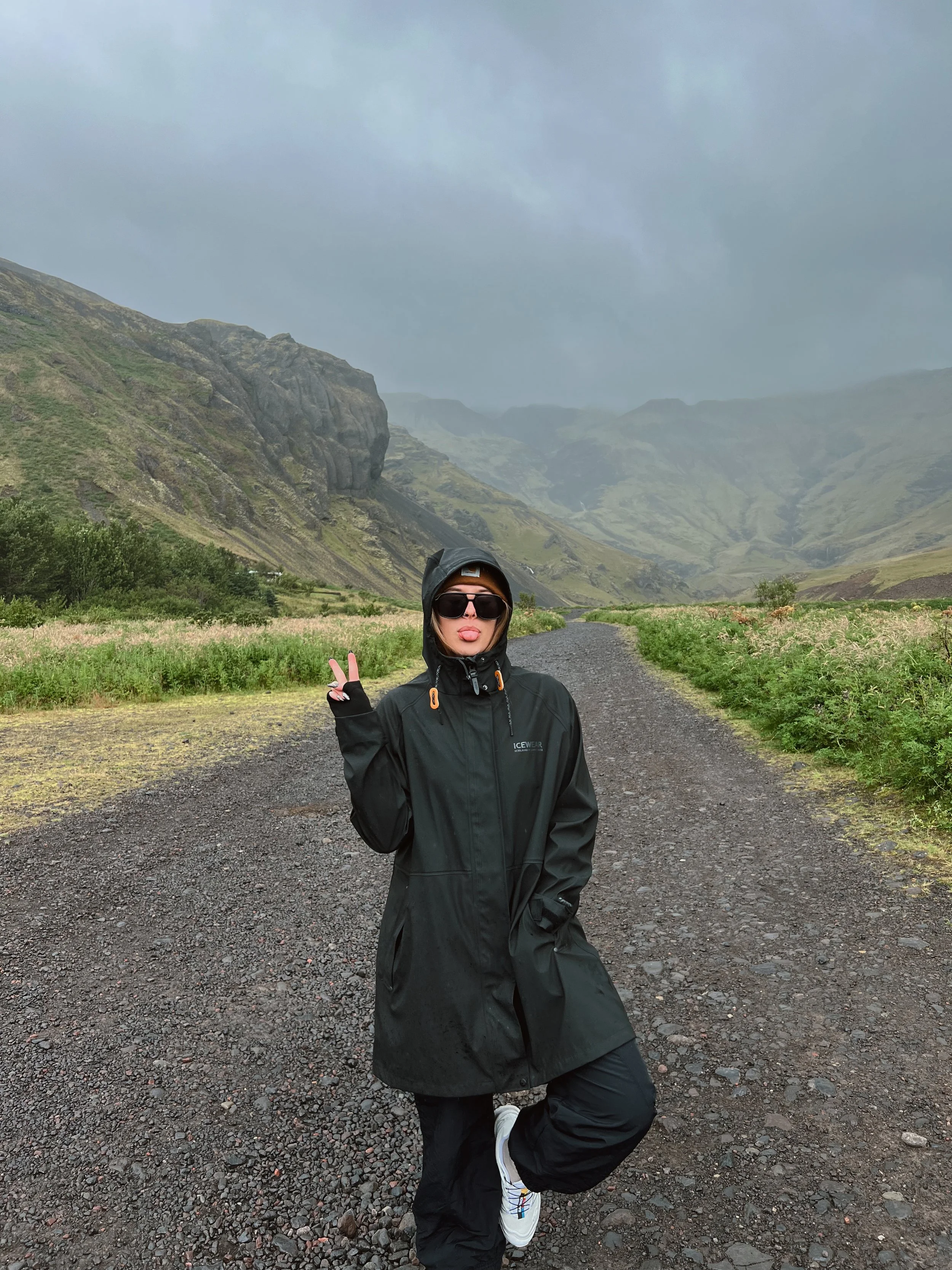 A woman wearing a black jacket, black pants, white sneakers, sunglasses, and a hood making a peace sign with her right hand while posing on a gravel trail in a mountainous landscape with overcast skies.