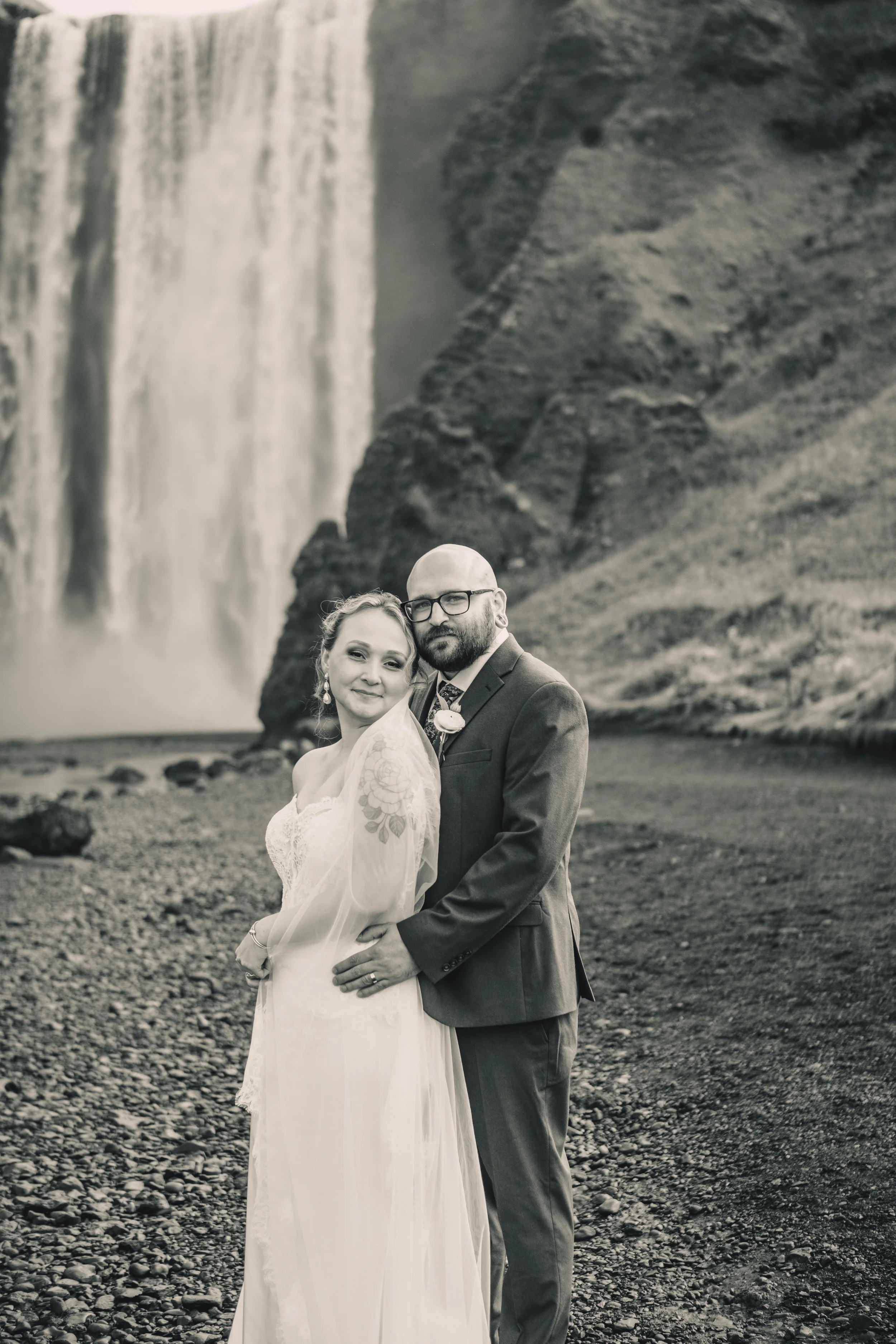 A black and white photo of a newlywed couple standing outdoors on a rocky terrain with a large waterfall and cliff in the background. The woman wears a flowing lace wedding dress and the man is dressed in a suit with glasses. They are close together,