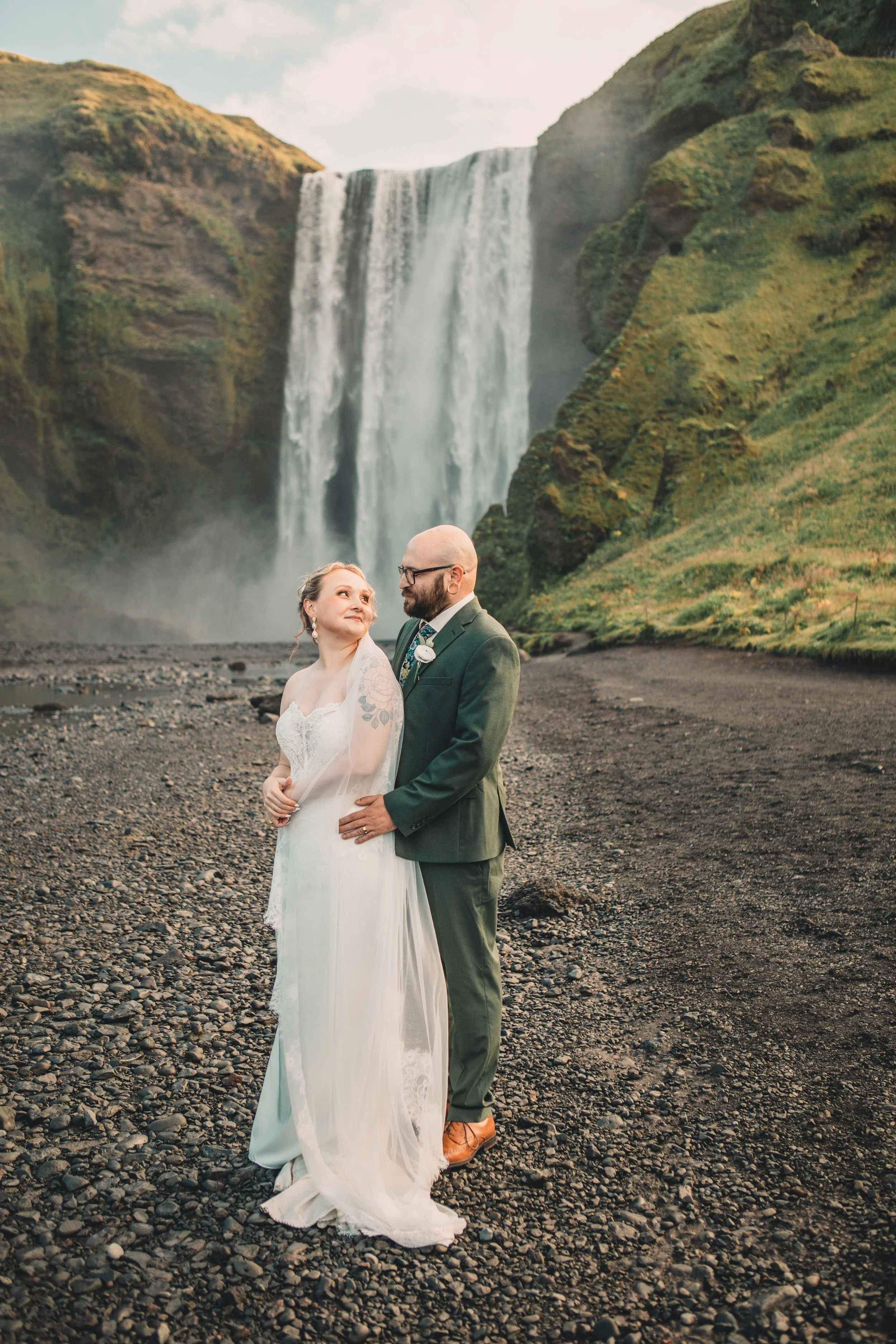 A bride and groom standing on a rocky beach in front of a large waterfall, with green hills on either side and a partly cloudy sky above.