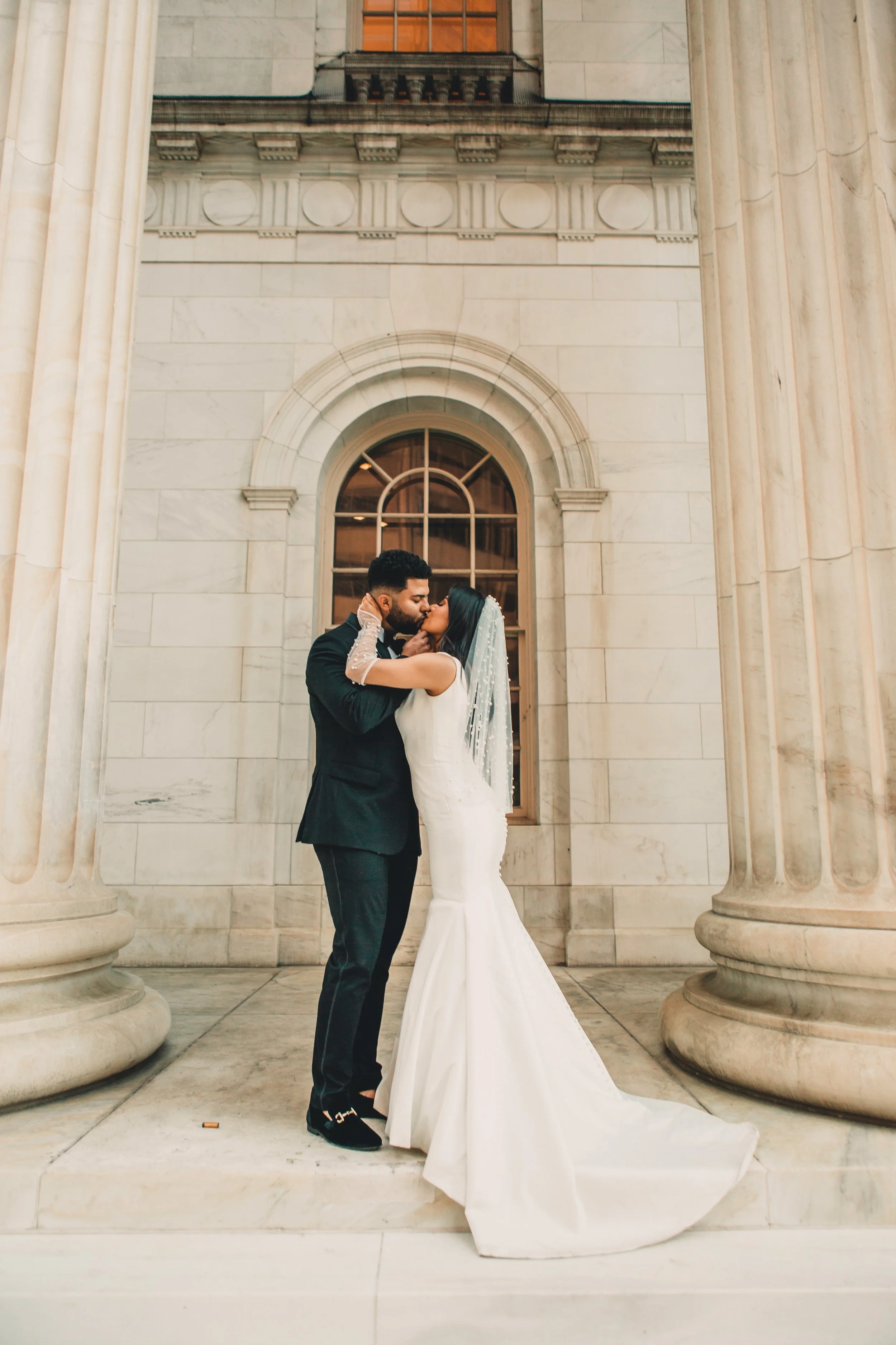 A bride and groom sharing a kiss, standing between large marble columns in front of a building with classical architecture. The bride wears a white gown and veil, and the groom is dressed in a dark suit.