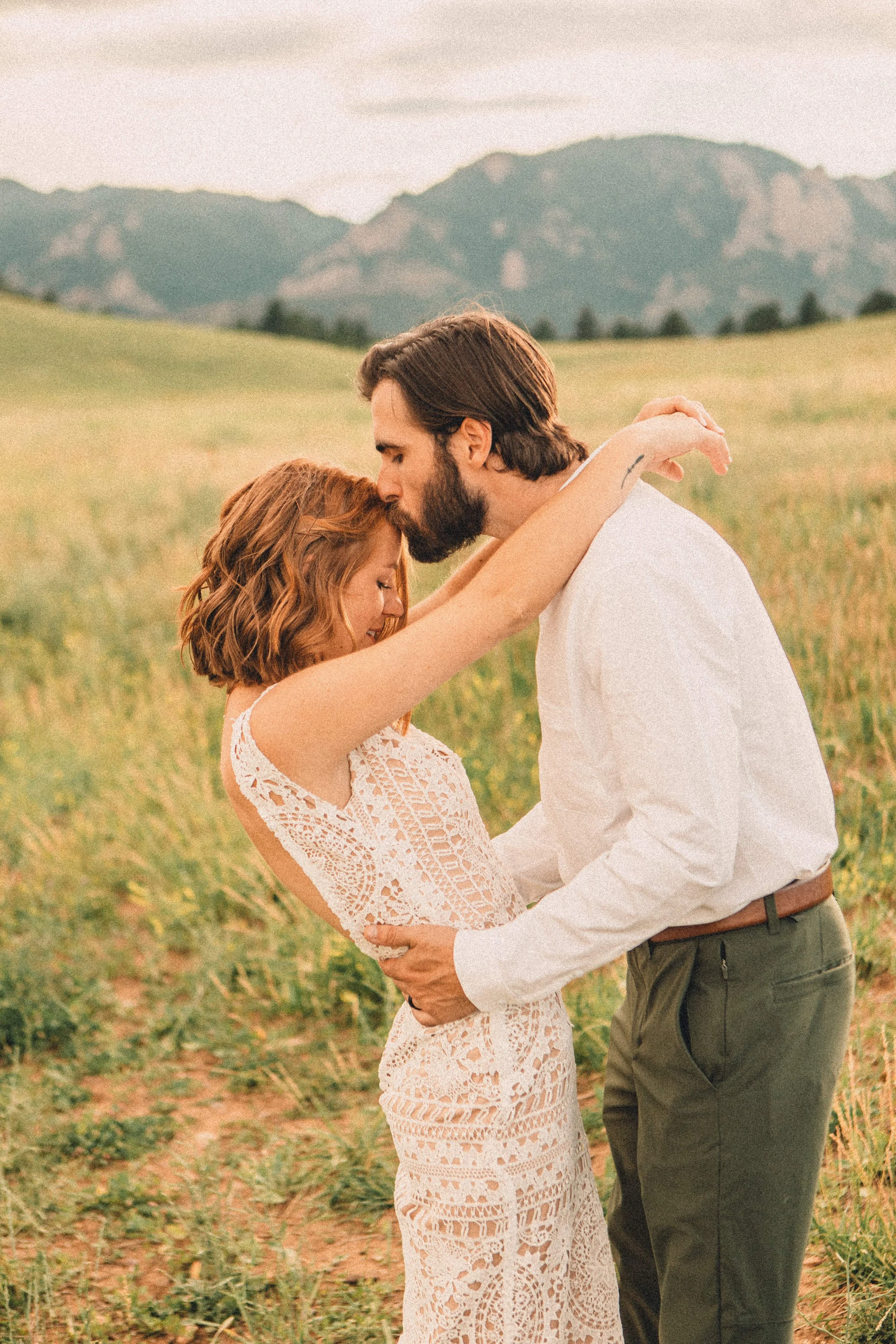A couple embraces in a grassy field with mountains in the background during sunset, with the man kissing the woman's forehead.