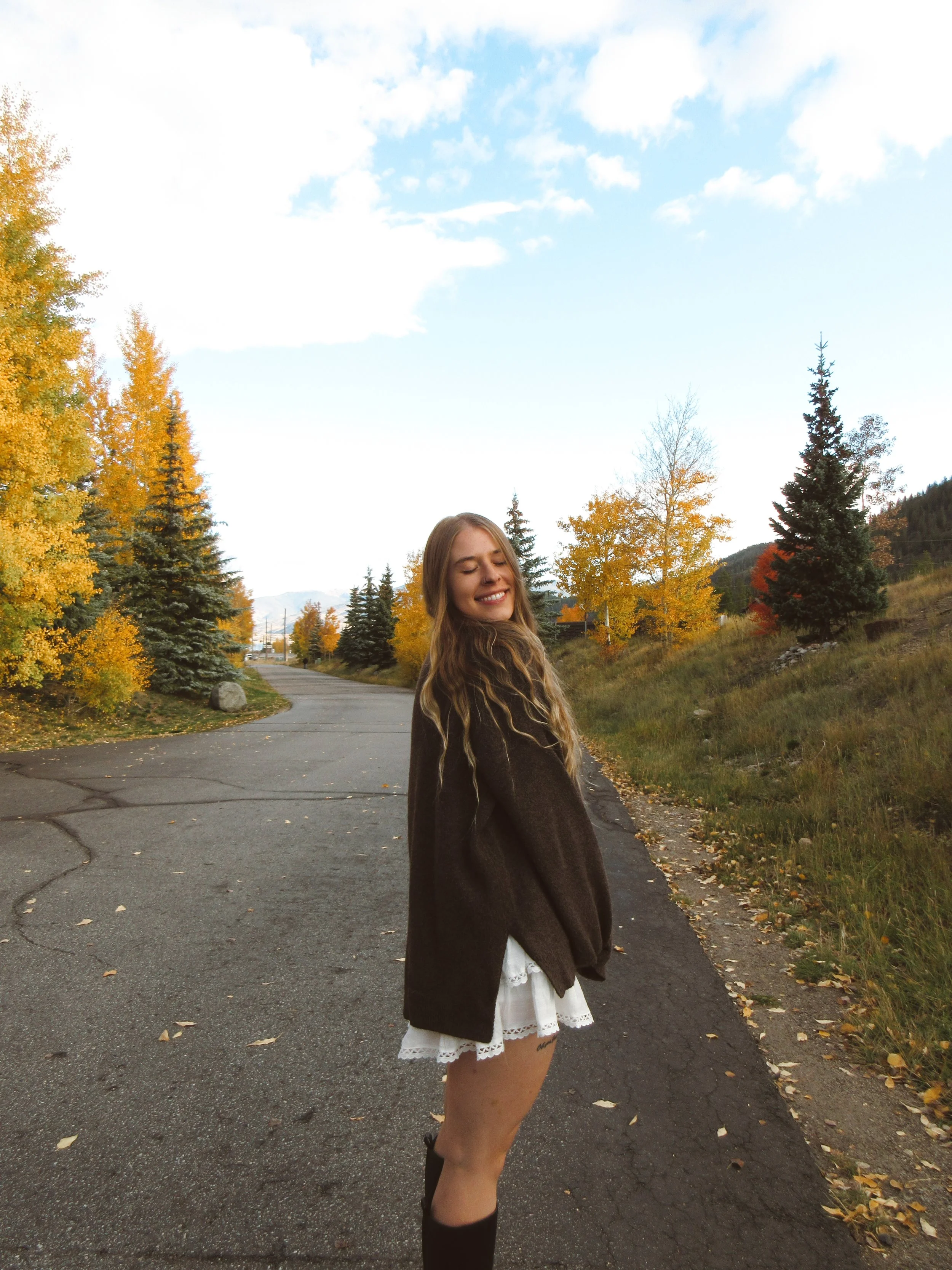 A young woman with long hair smiling with eyes closed, standing on a paved outdoor path with autumn trees and mountains in the background.
