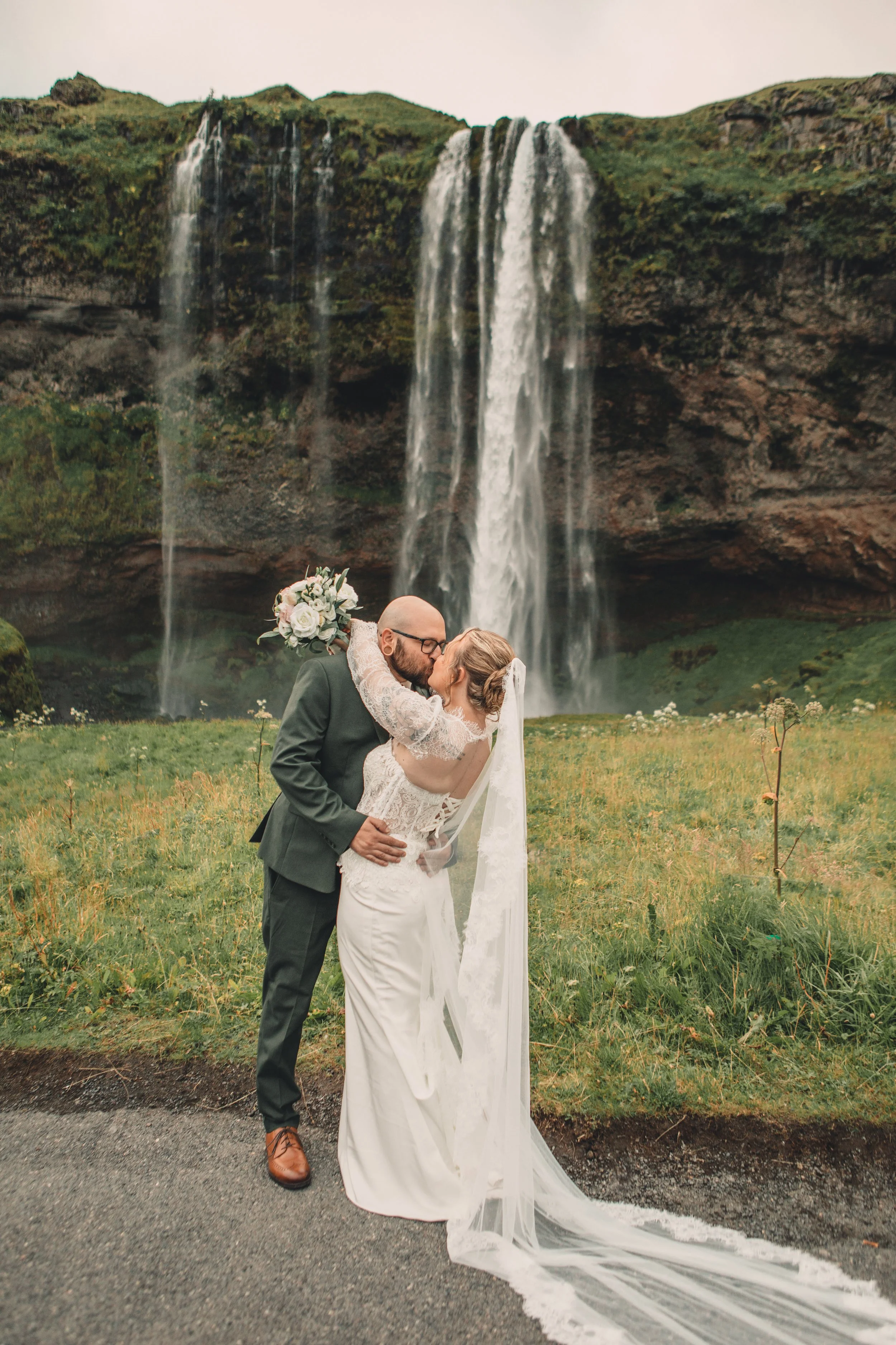 A bride and groom kissing outdoors in front of a waterfall, with the bride wearing a lace wedding dress and holding a bouquet, and the groom in a dark suit and brown shoes.
