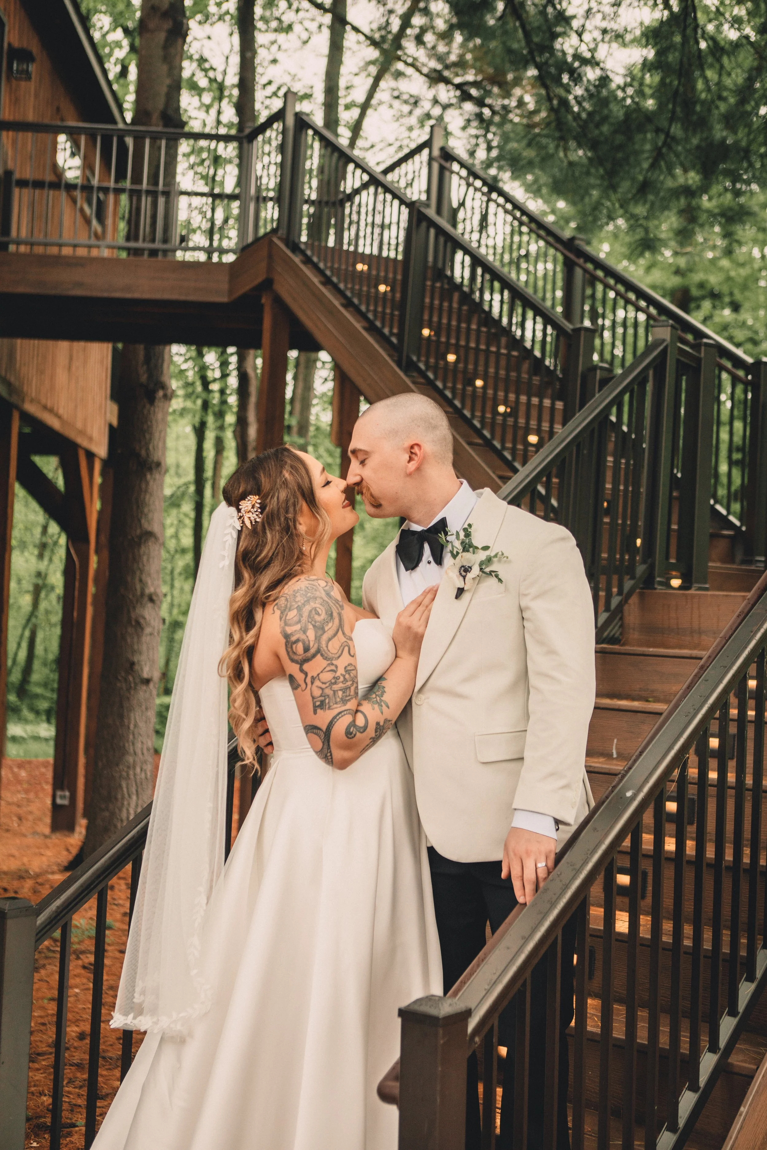 A bride and groom sharing a kiss outdoors under a staircase surrounded by trees during their wedding.