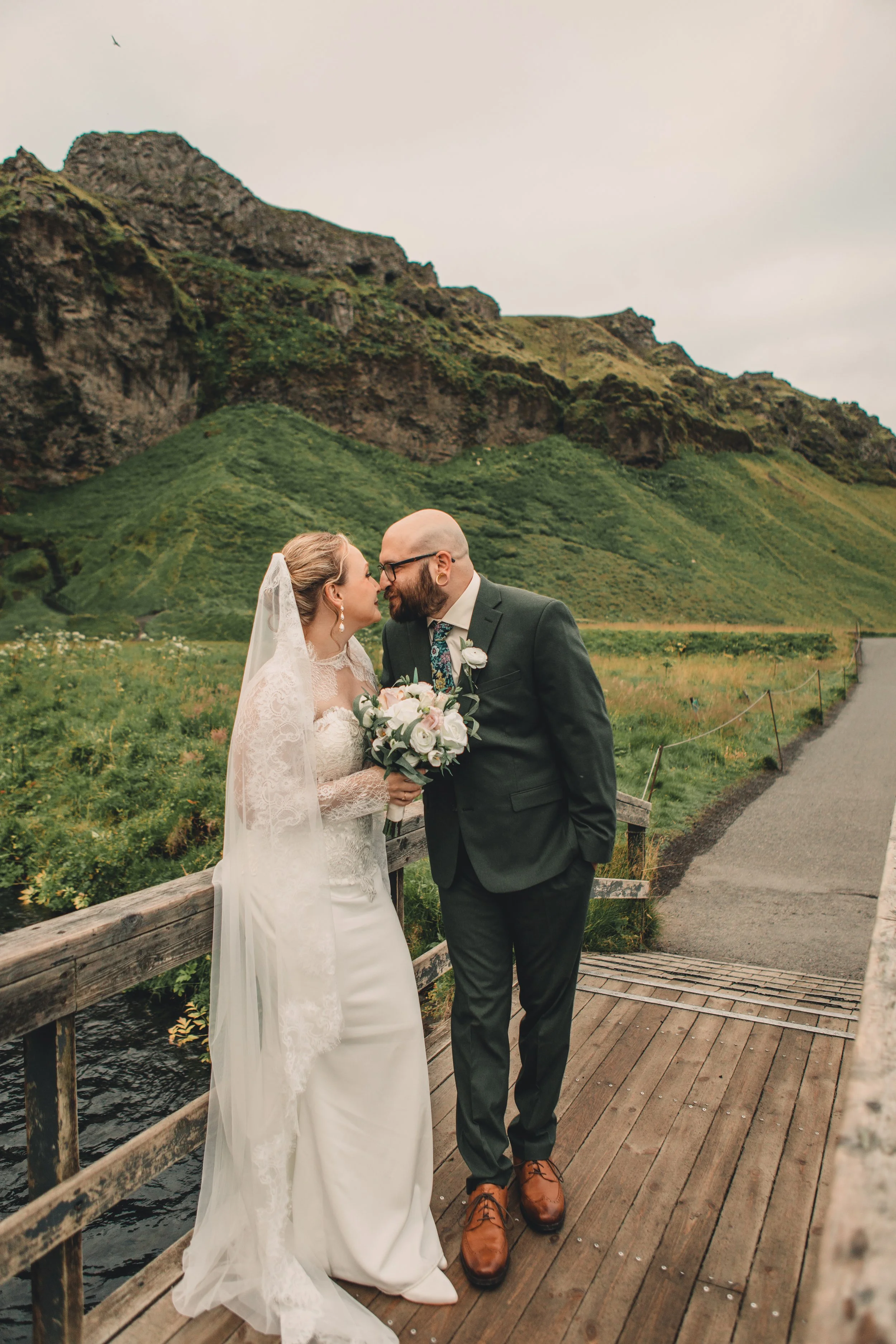 A bride and groom standing on a wooden bridge with a scenic mountainous landscape in the background, sharing a close moment.