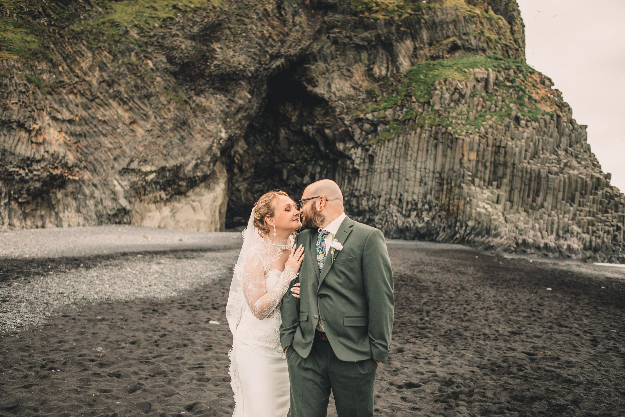 a newlywed couple standing on a rocky beach with towering volcanic cliffs behind them, sharing a kiss and holding each other in a romantic pose.