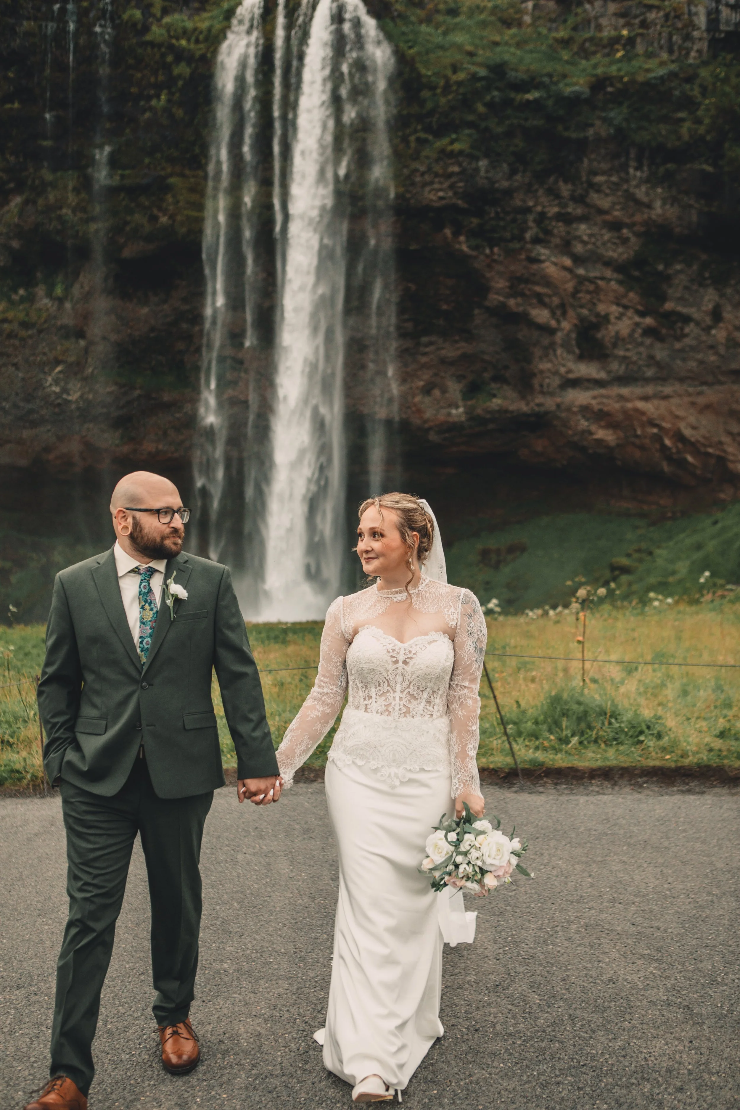 A couple in wedding attire holding hands, walking outdoors with a waterfall in the background. The groom, on the left, is wearing a dark suit, white shirt, and a colorful tie. The bride, on the right, is wearing a lace wedding dress, holding a bouque