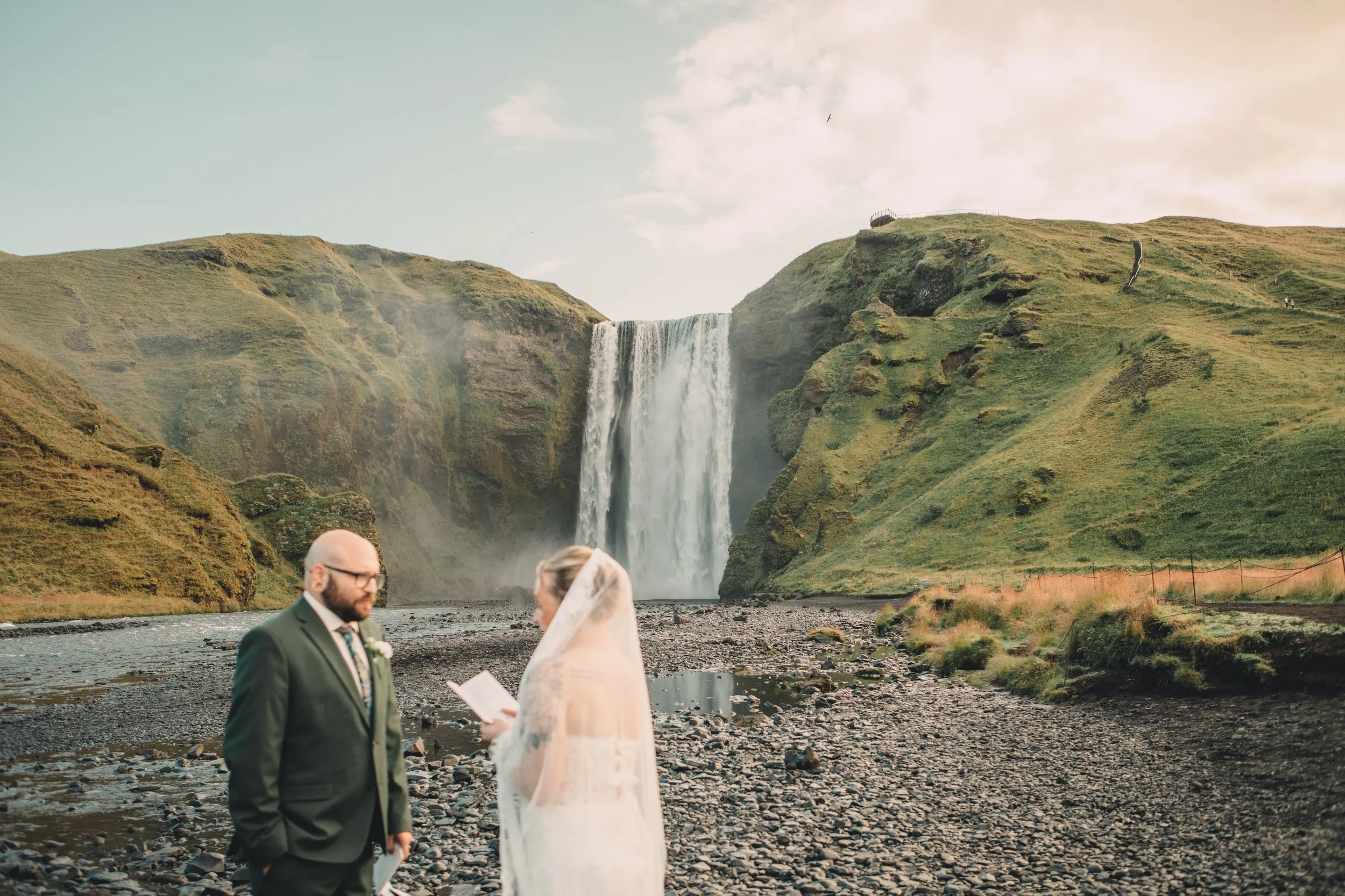 A bride and groom are standing outdoors near a river, having a wedding ceremony, with a large waterfall and green hills in the background.