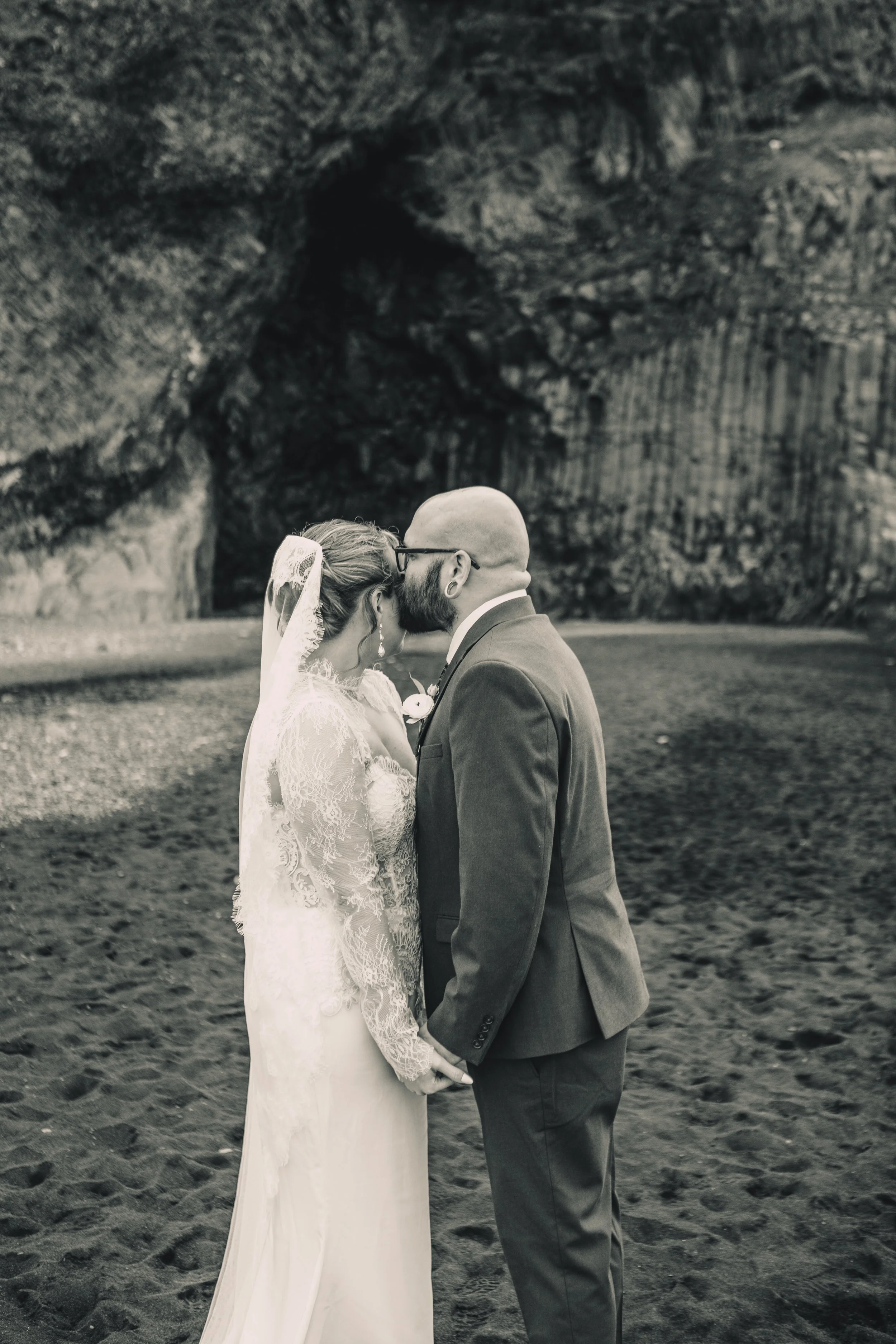 A bride and groom stand close together holding hands and sharing a kiss in front of a natural cave or cliff formation.