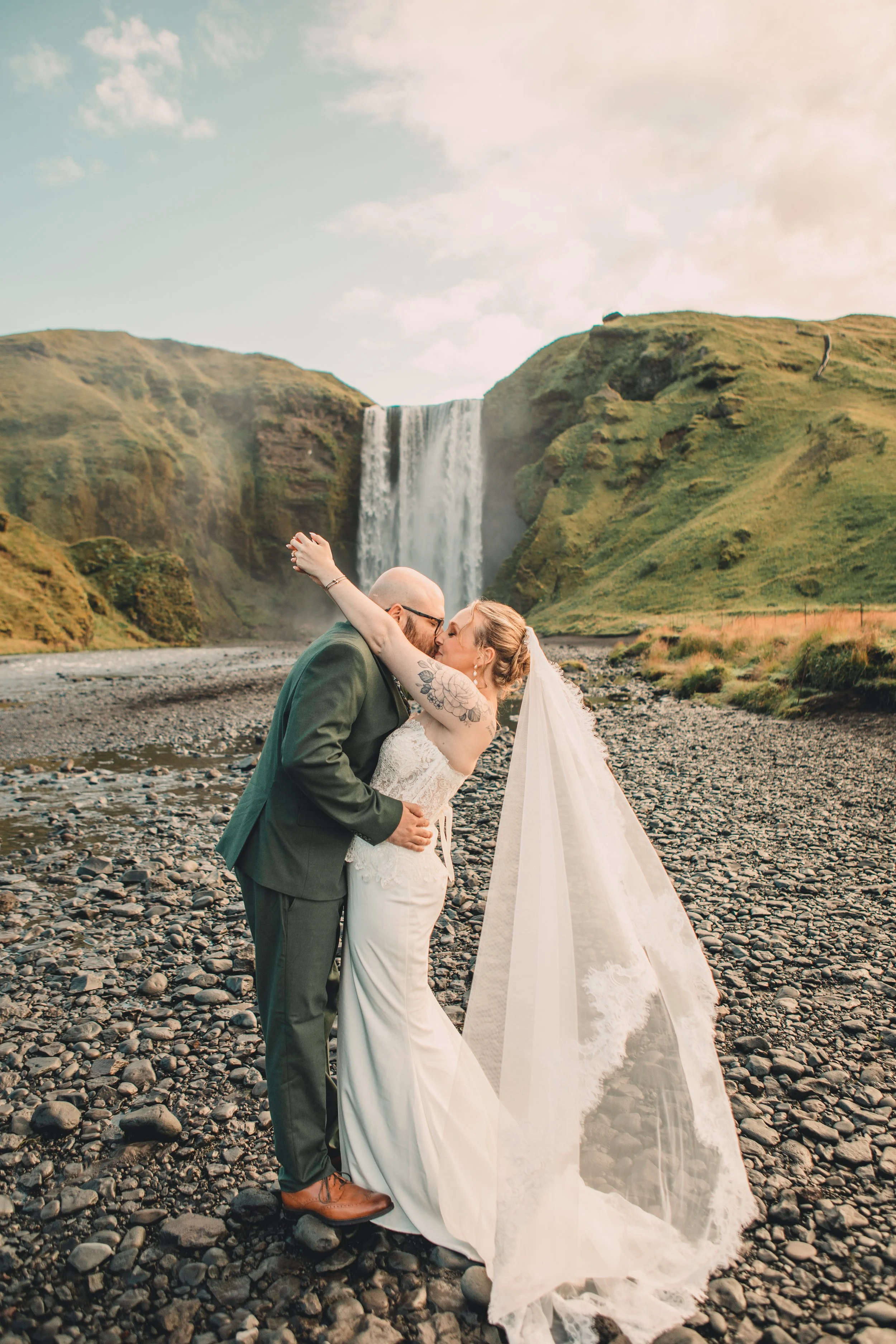 A newlywed couple kissing on a rocky riverbed with a waterfall and green hills in the background.
