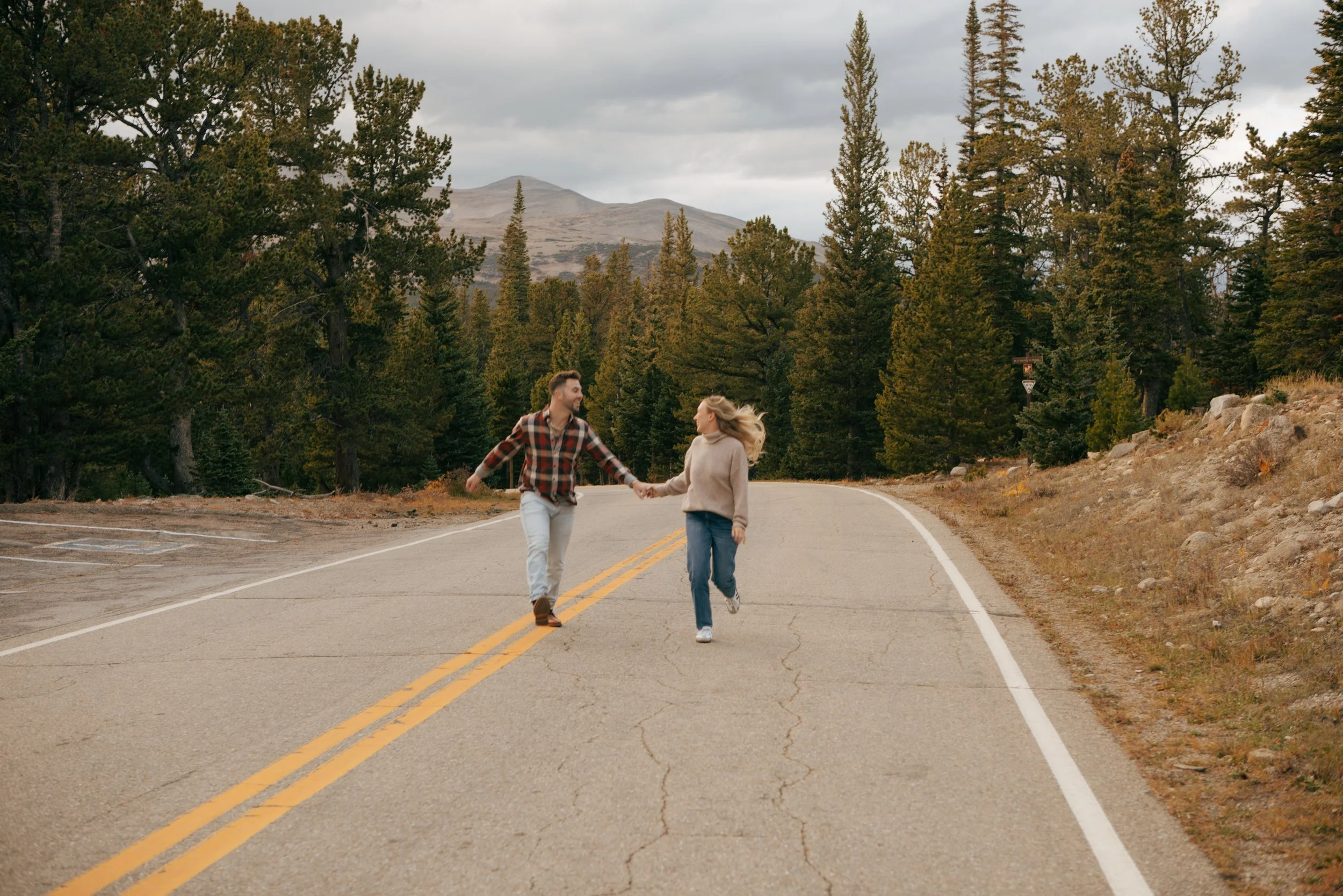 A couple holding hands and running on a winding mountain road surrounded by tall pine trees, with mountains in the background and a cloudy sky overhead.