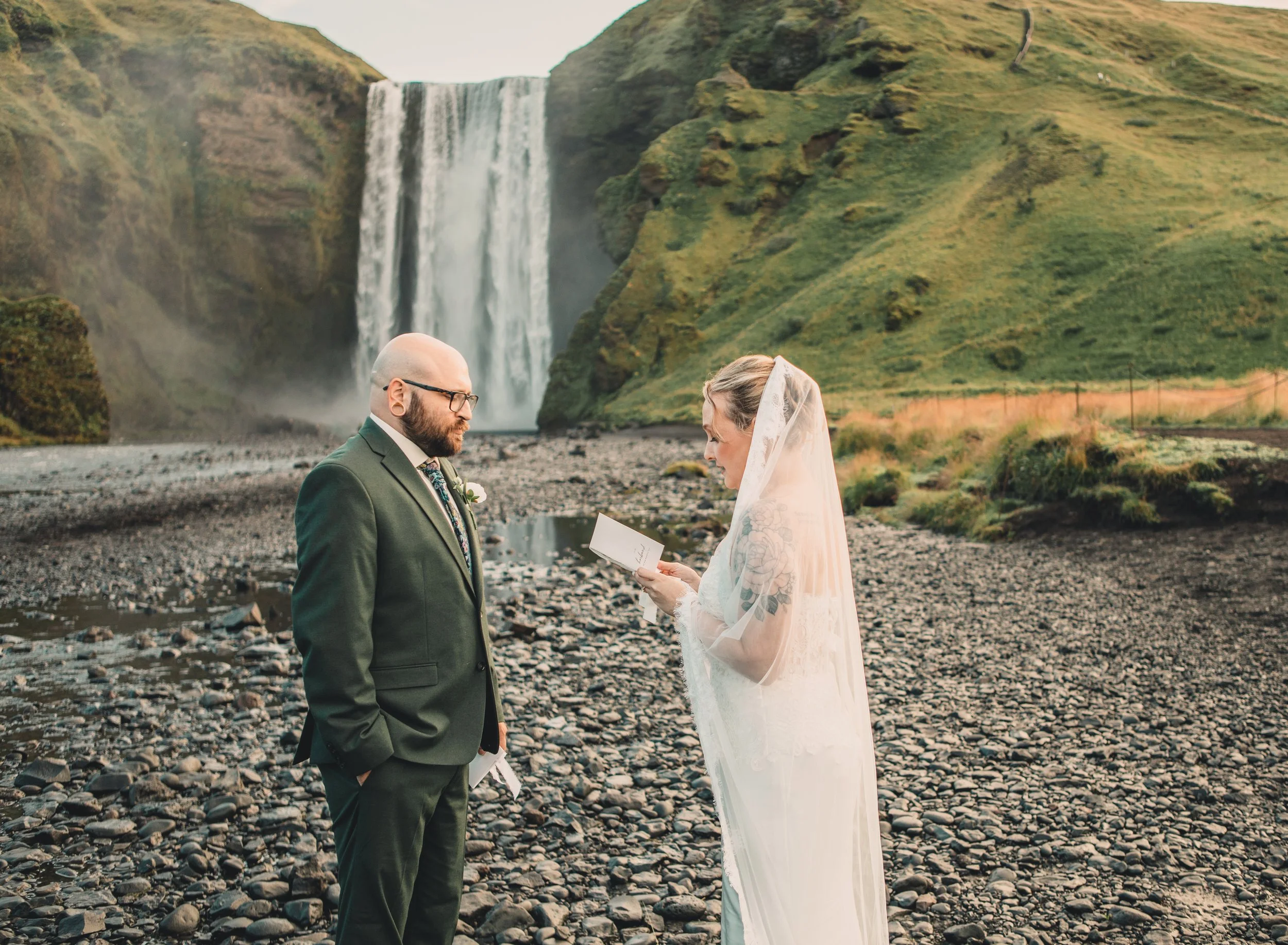 A couple is standing outdoors near a river with a waterfall in the background, holding a piece of paper, likely during a wedding or vow exchange. The man is wearing a dark suit and glasses, and the woman is wearing a lace wedding dress with a veil, r