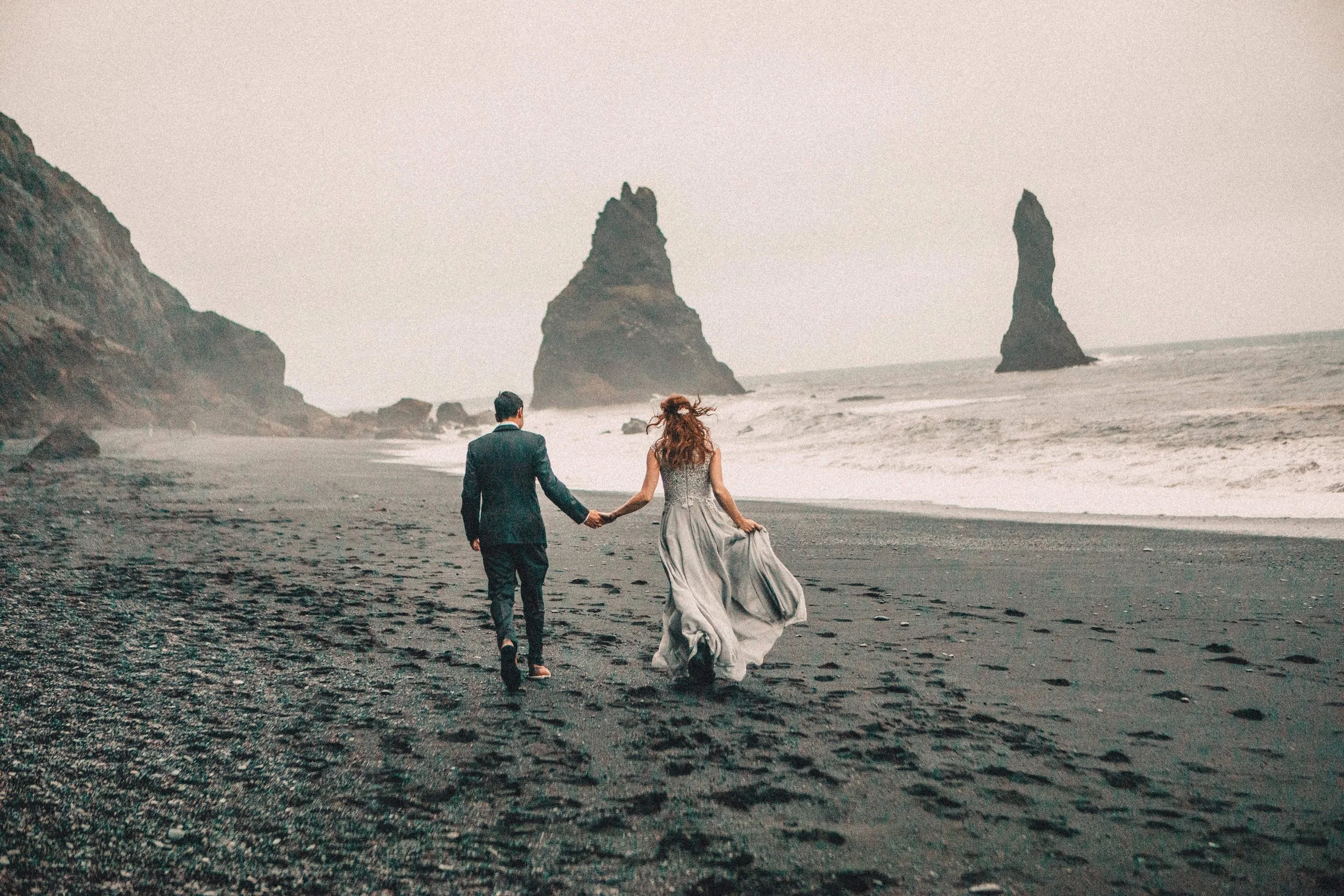 A couple dressed in wedding attire walking hand in hand on a black sand beach with large rock formations and cliffs in the background.