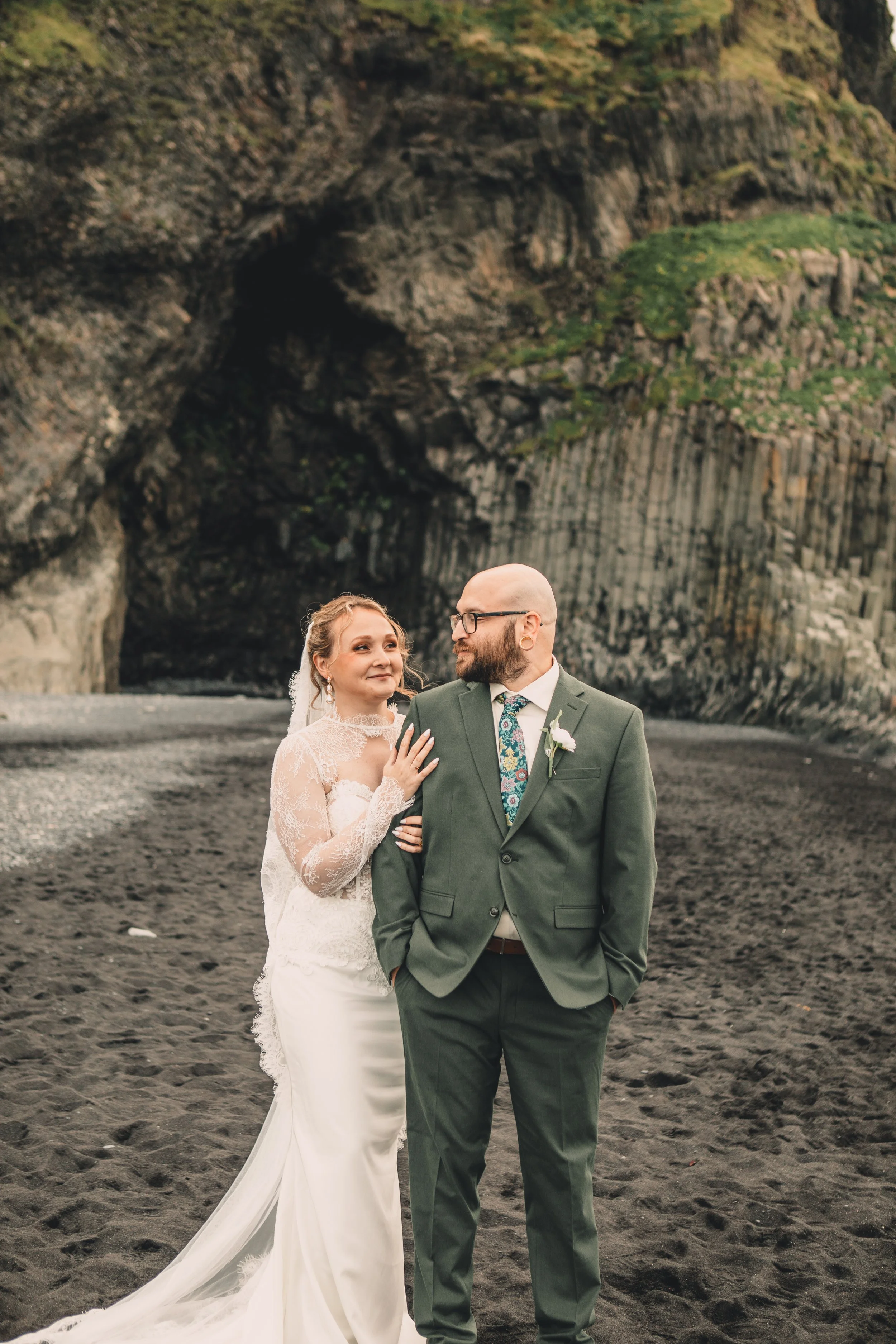 A bride and groom standing on a black sand beach with a large cliff and cave in the background. The bride is wearing a long white lace wedding gown with a veil, and the groom is wearing a green suit with a colorful tie and glasses. They are looking at each other affectionately.
