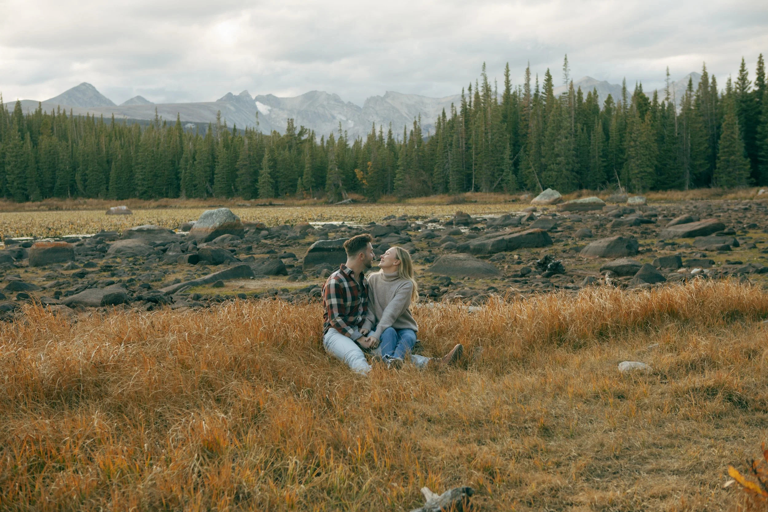 A couple sitting closely together in a field of orange grass, with a backdrop of a forest of pine trees and mountains under a cloudy sky.
