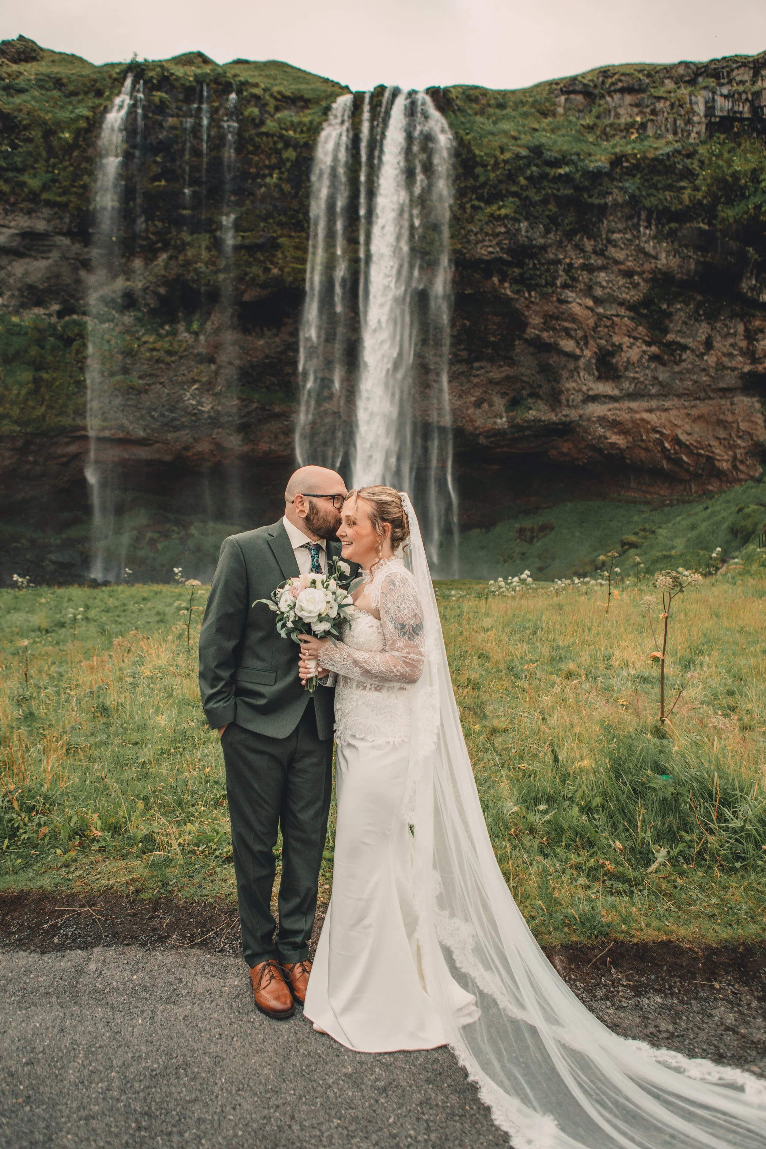 A bride and groom kissing in front of a large waterfall during a wedding photoshoot. The bride is holding a bouquet and wearing a long white lace wedding dress with a train, and the groom is wearing a dark suit with a patterned tie and glasses.