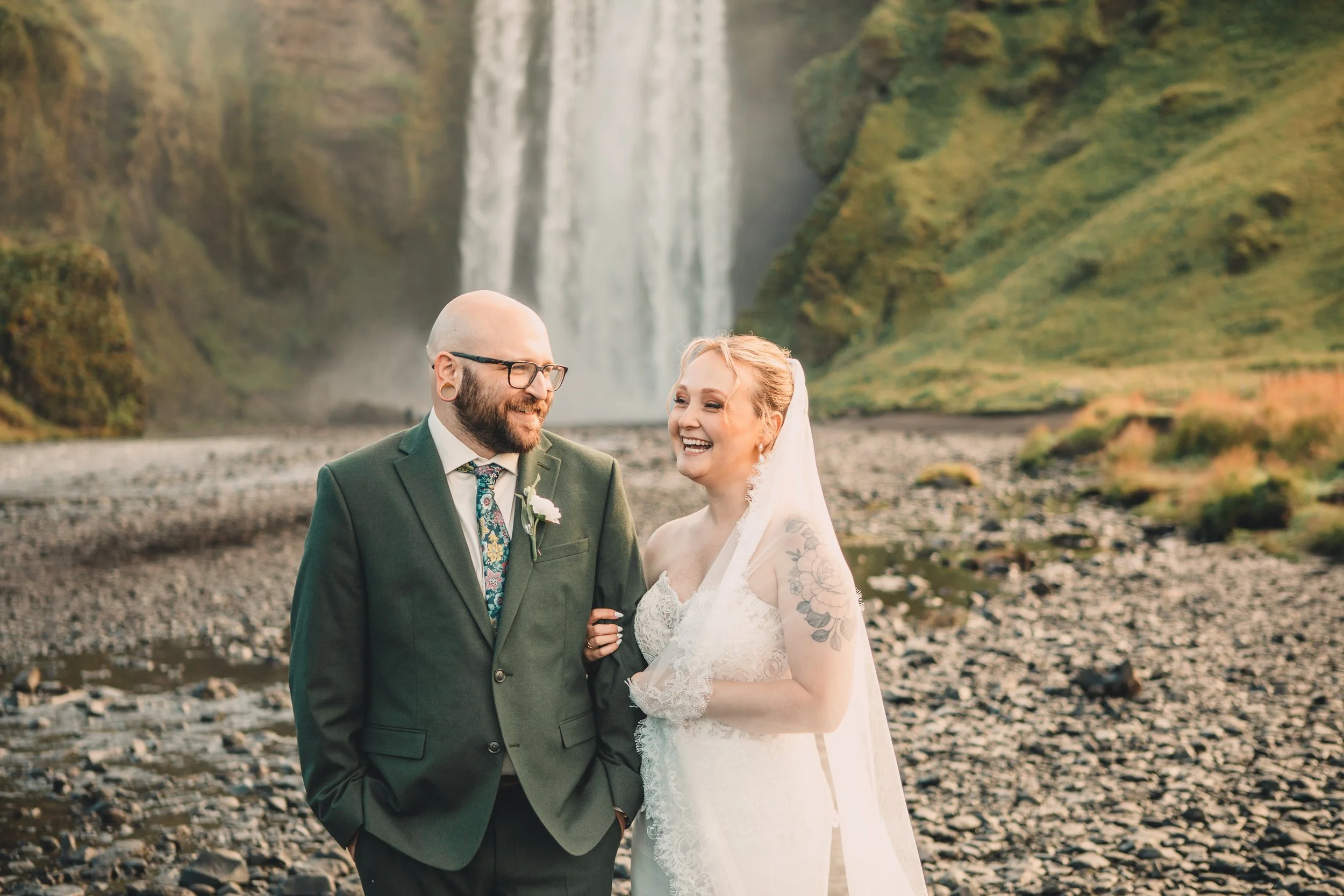 A smiling bride and groom stand outdoors near a waterfall, holding hands. The groom wears glasses and a dark suit, and the bride has a white lace dress and visible tattoos on her arm. The background features a large waterfall cascading down a green, moss-covered cliff.