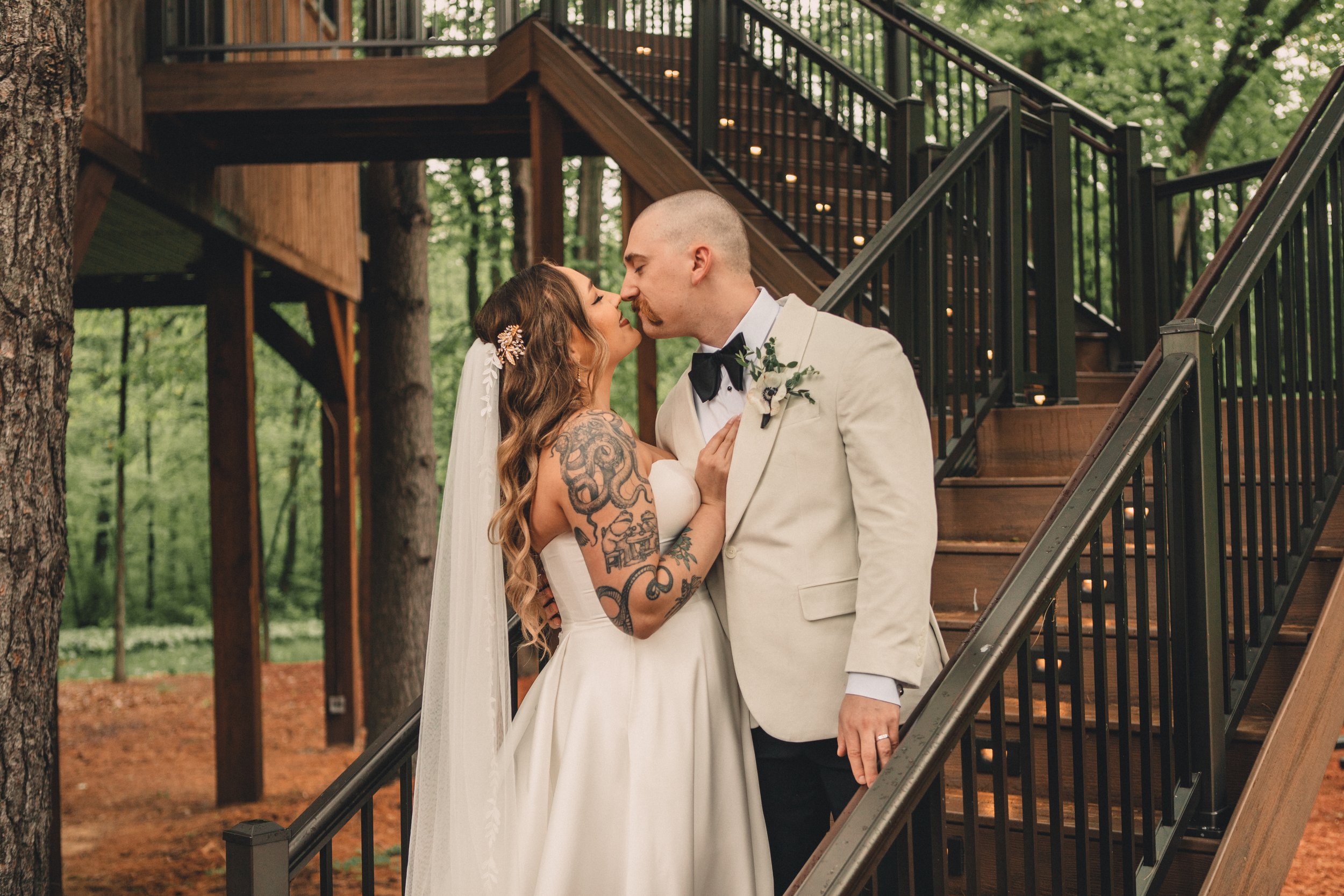 A bride and groom sharing a kiss outdoors near a wooden staircase in a wooded area.