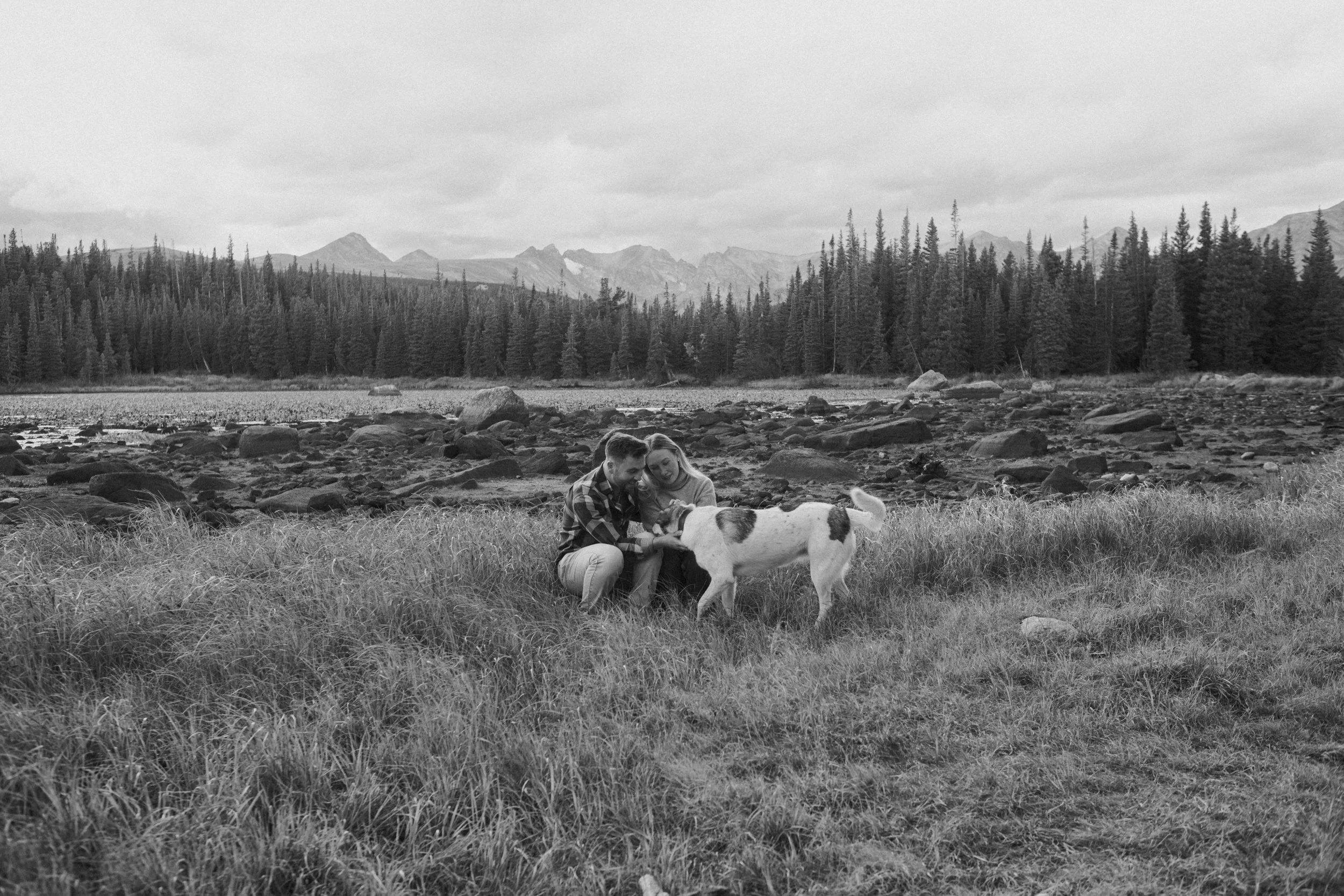 A couple sits on grass near a river, smiling at a white dog with dark spots. Behind them is a forest of tall trees, mountains, and an overcast sky.