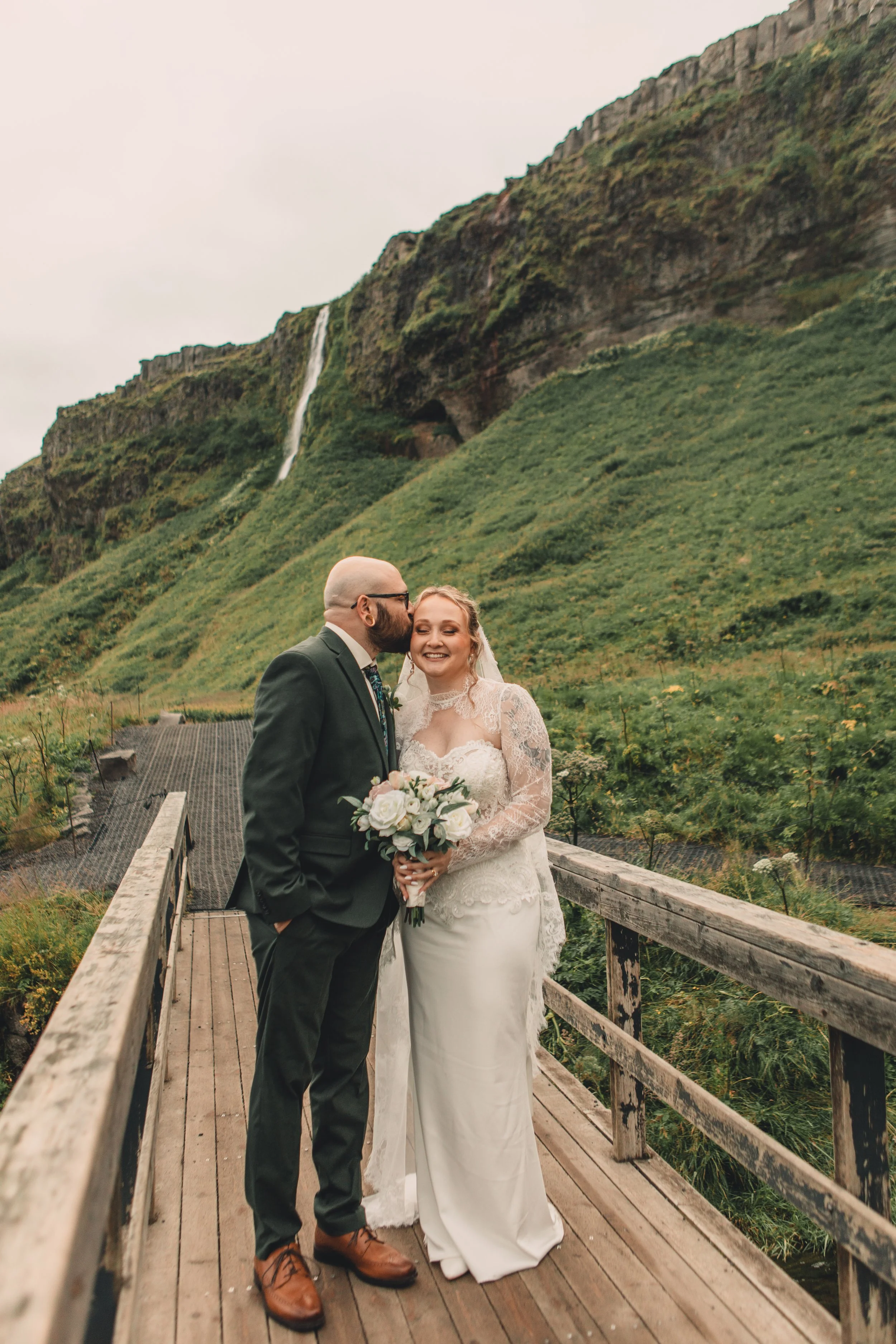A newlywed couple standing on a wooden bridge outdoors, with the groom kissing the bride's temple. The bride is holding a bouquet of white roses. The background features lush green hills and a waterfall.