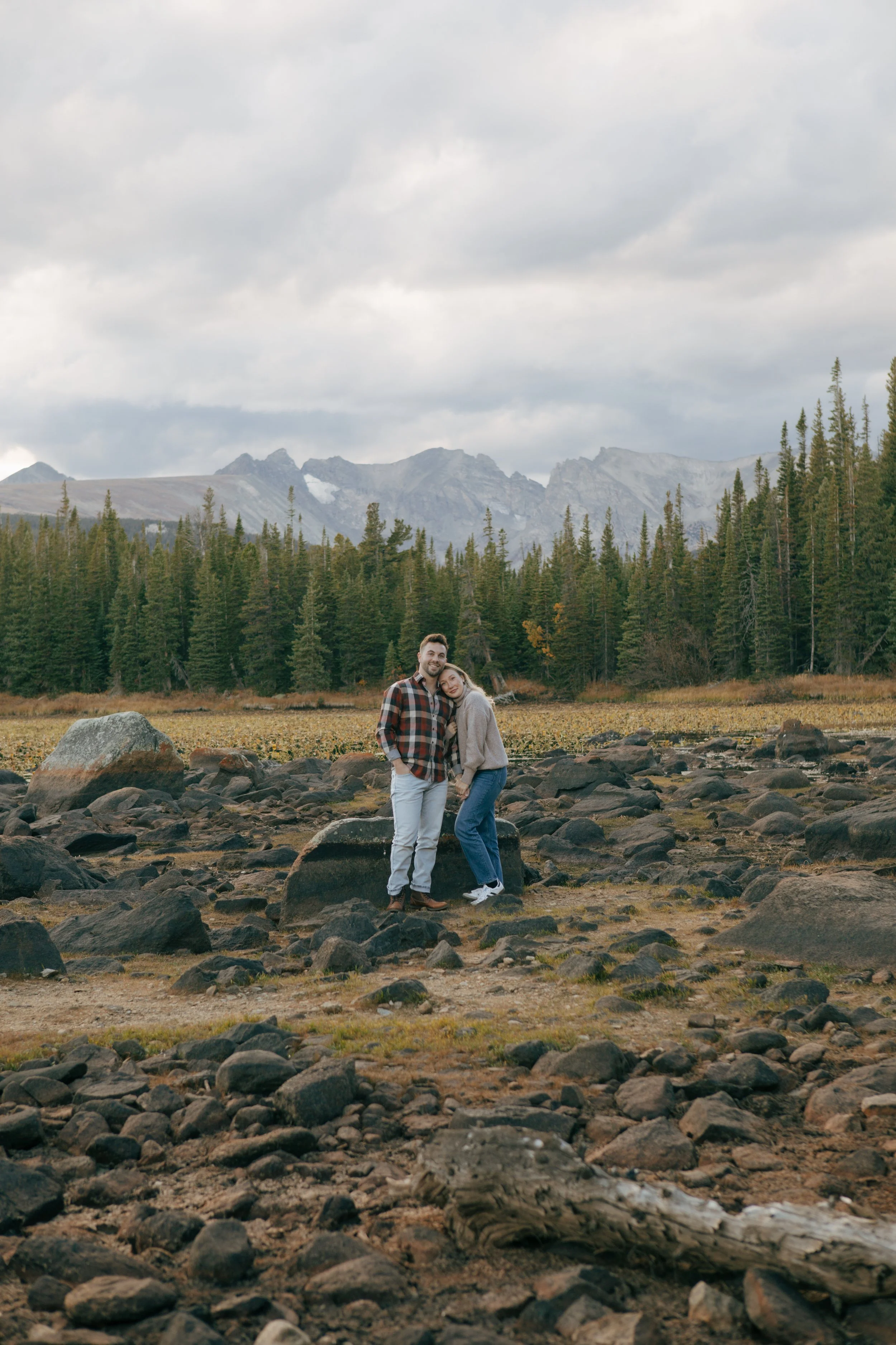 A couple standing on rocks in a valley with a mountain range and cloudy sky in the background.