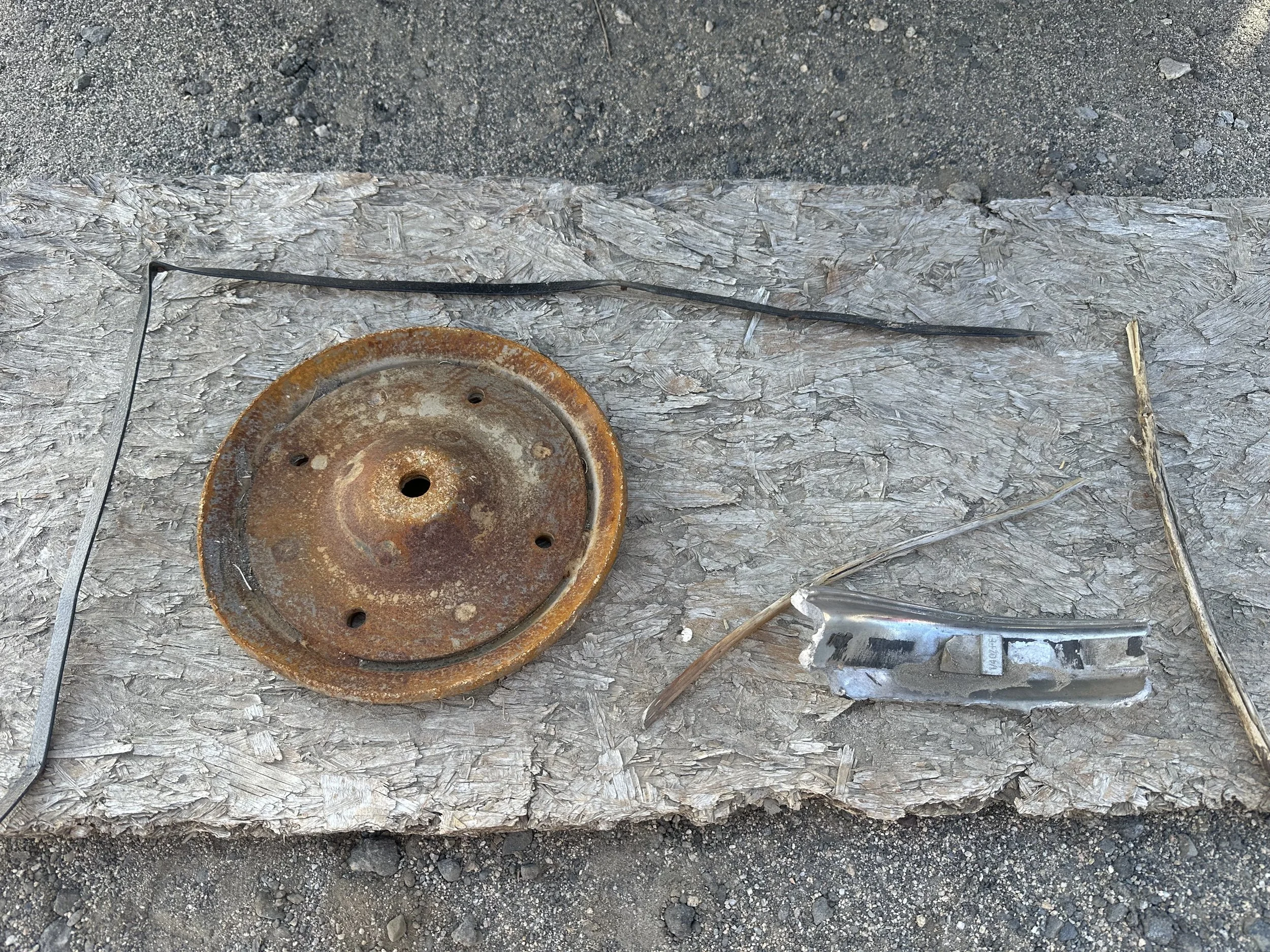 A weathered OSB plywood on gravel with rusty steel, metal, a piece of an aluminum wheel.