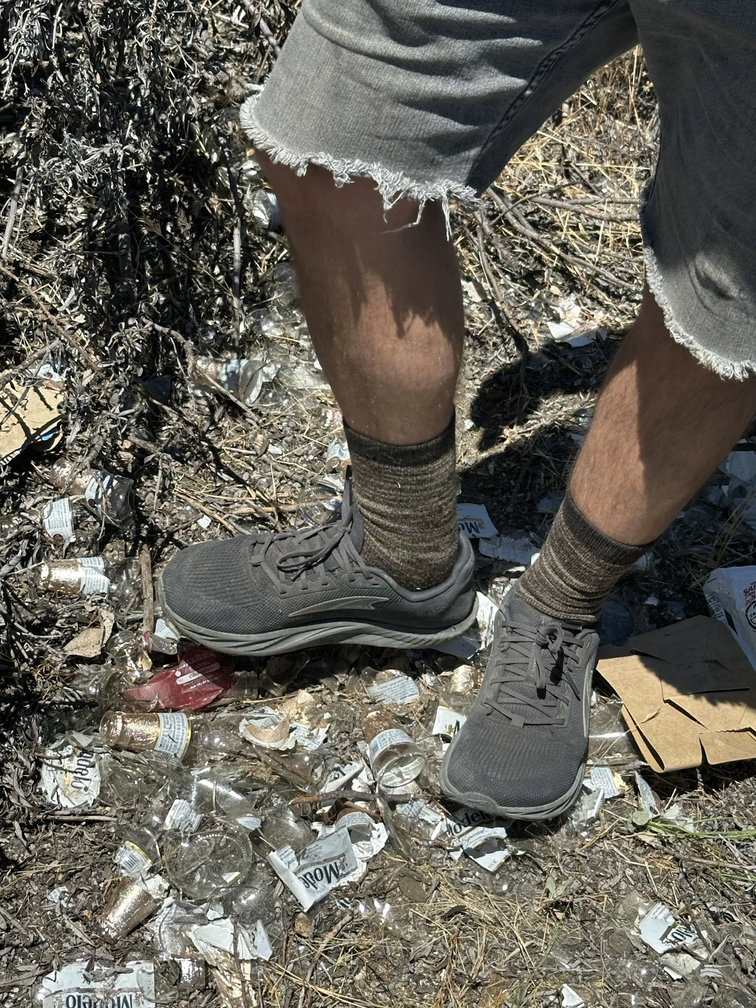 Legs wearing cut off black jeans, brown socks and black shoes steps on Modelo beer glass and cardboard.