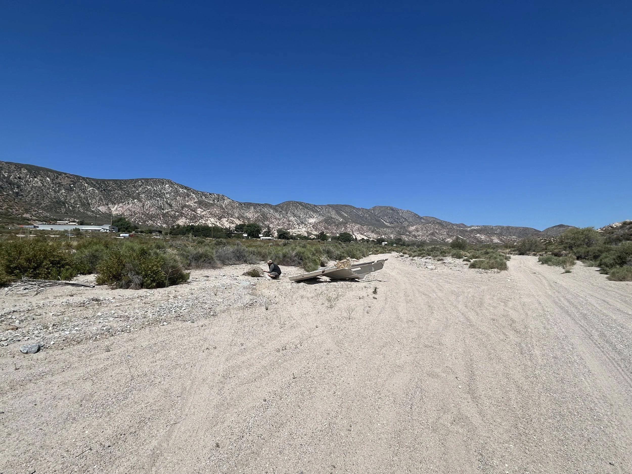 on s dirt road with bushes, cars, mountains and a big blue sky. A person is recording with a damaged metal boat behind them.