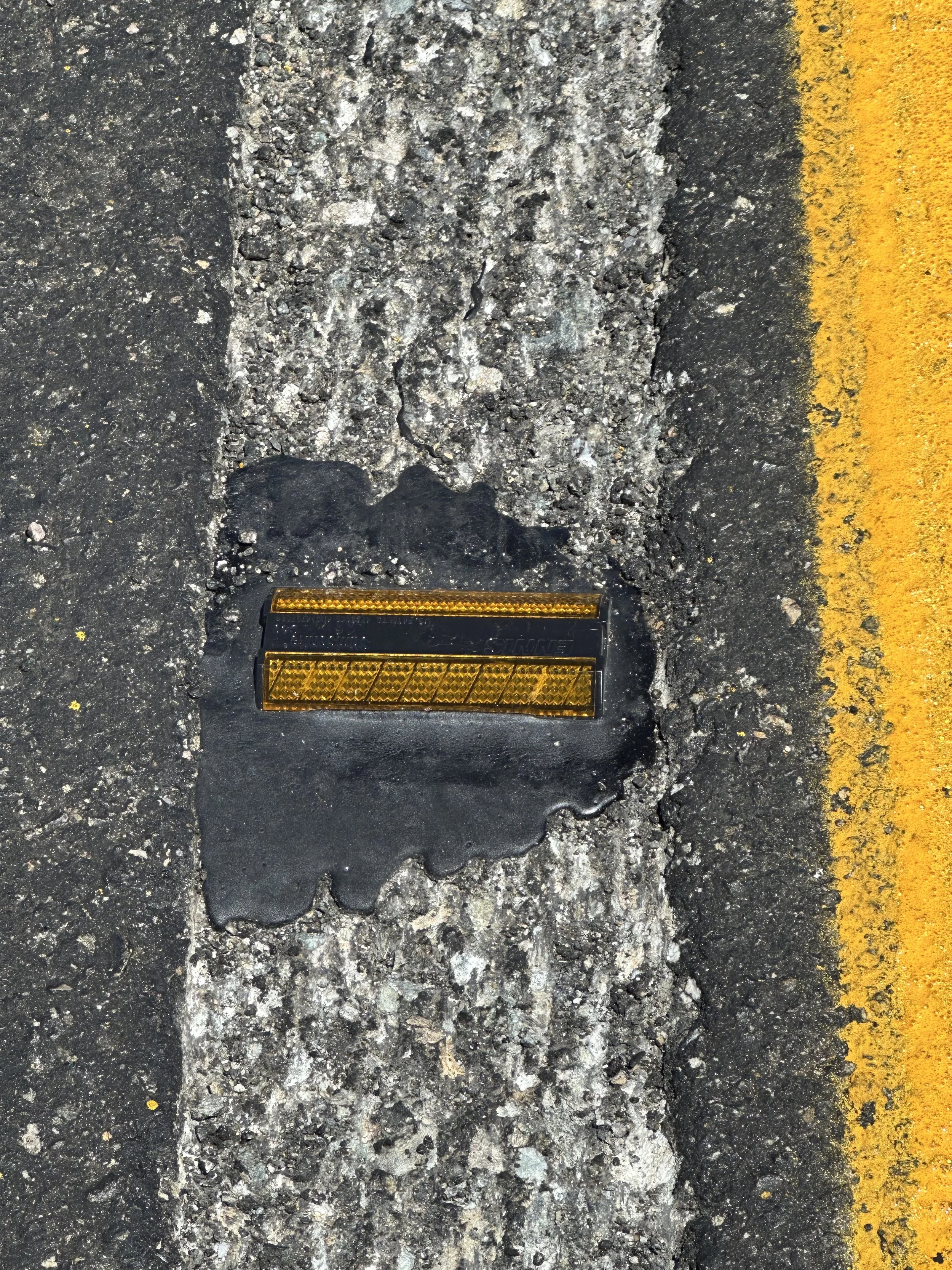 A yellow acrylic rumble bump installed with tar, on a white road paint strip, on asphalt.