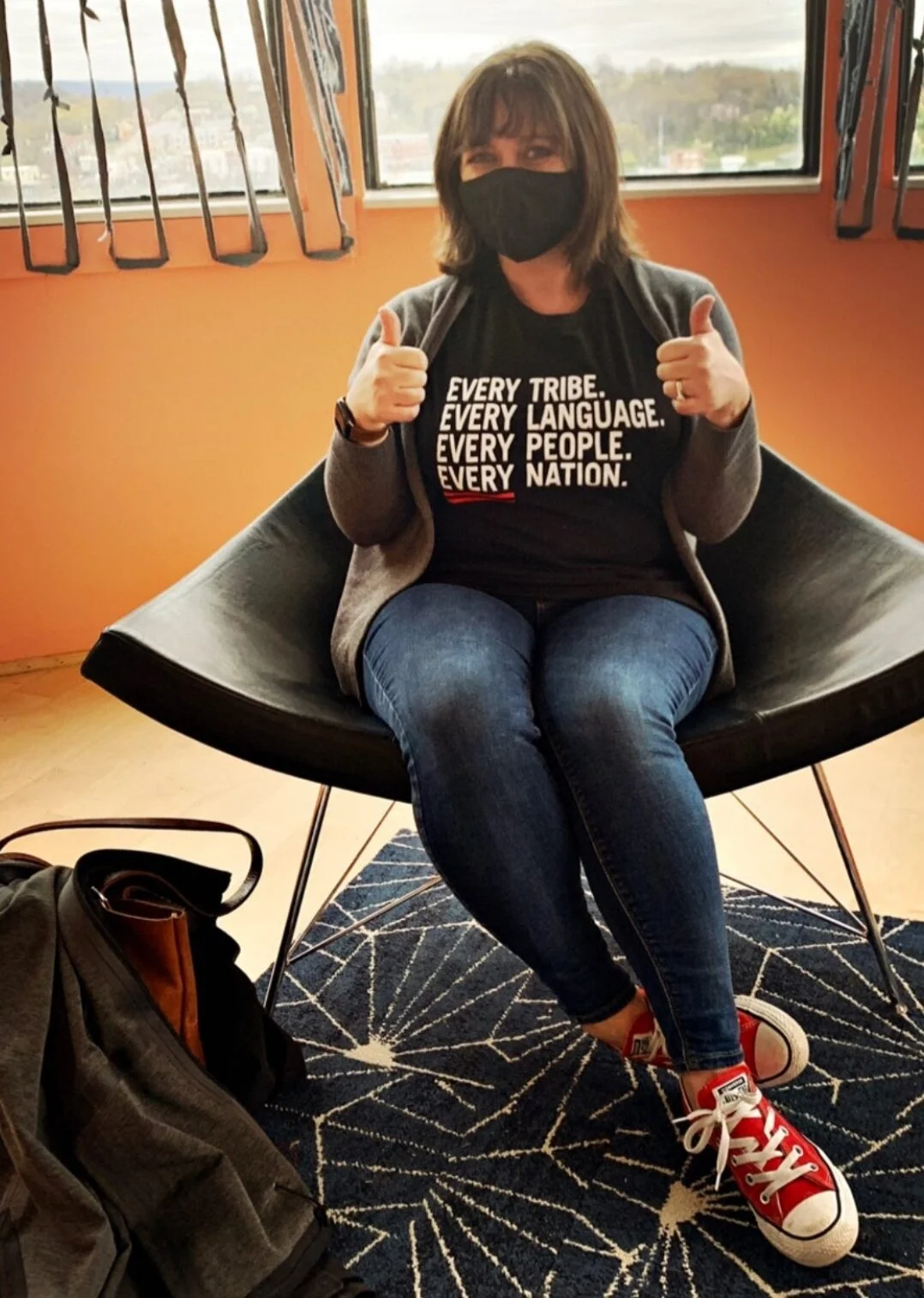 Person sitting in a chair wearing a mask, t-shirt with text "Every Tribe. Every Language. Every People. Every Nation.", jeans, and red sneakers, giving thumbs up.