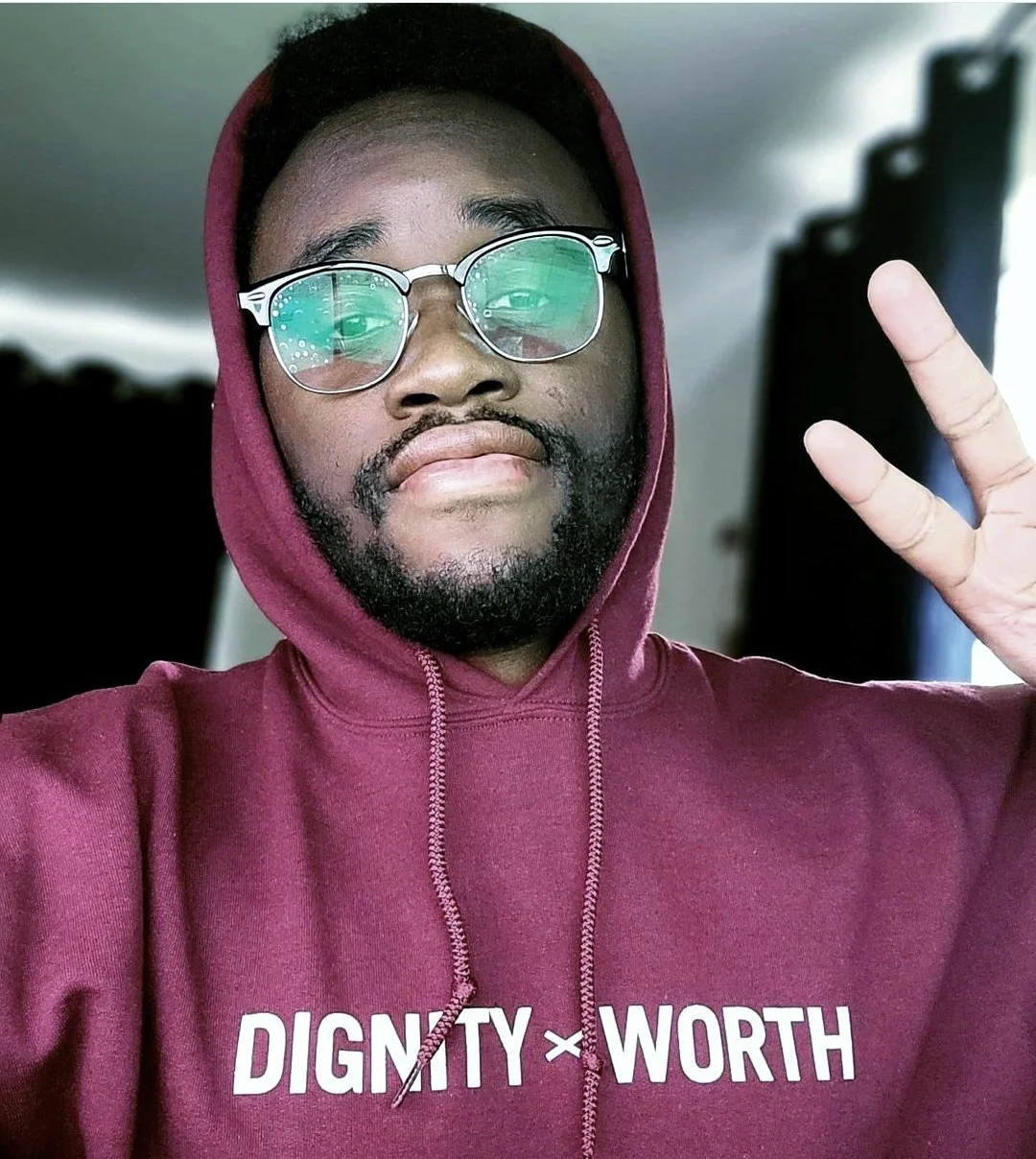 Person wearing glasses and a maroon hoodie with the words "DIGNITY x WORTH," making a peace sign gesture.