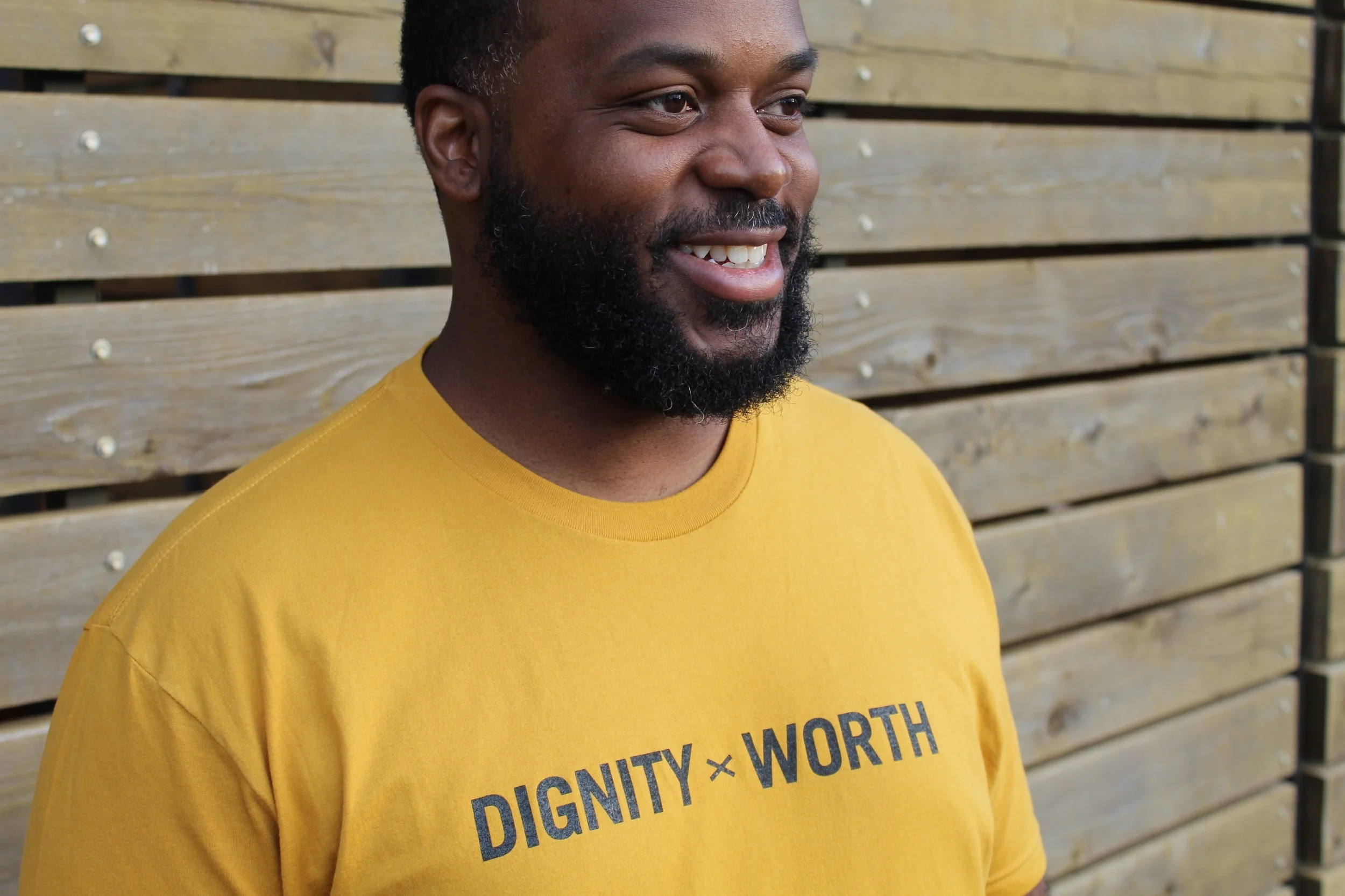 Man in a yellow t-shirt with text "Dignity x Worth" standing in front of a wooden wall, smiling.